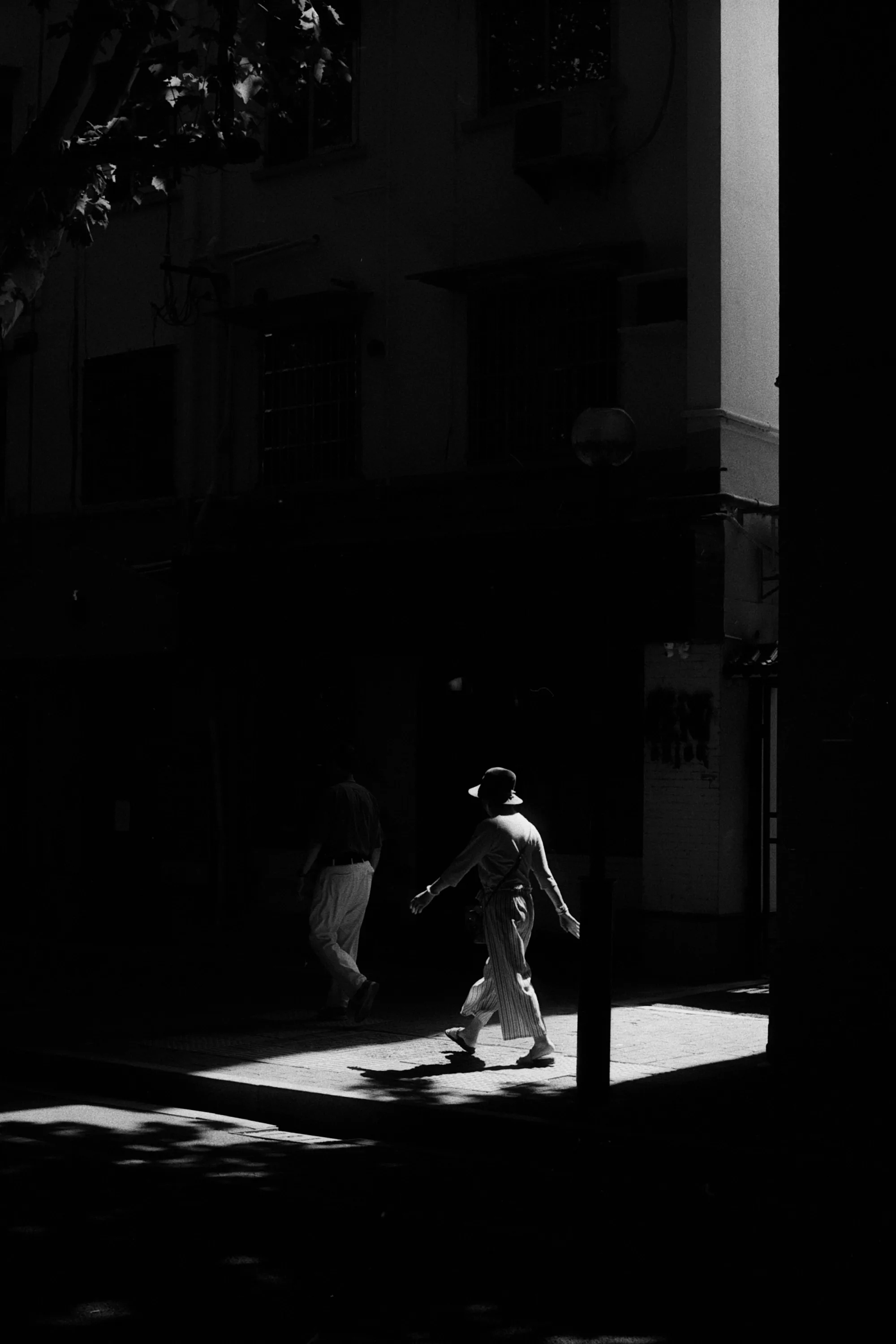 A lone pedestrian crosses a bright patch of pavement between deep building shadows.