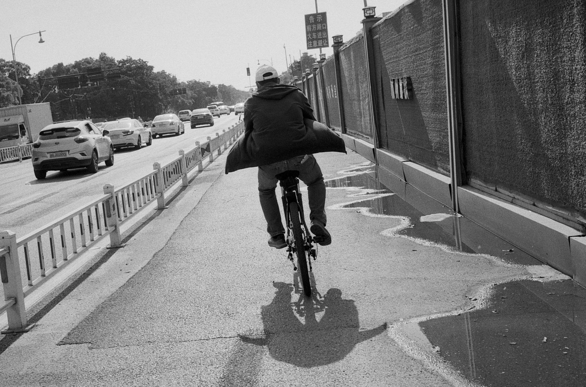 A cyclist rides alone along a narrow lane between traffic barriers and a construction fence.
