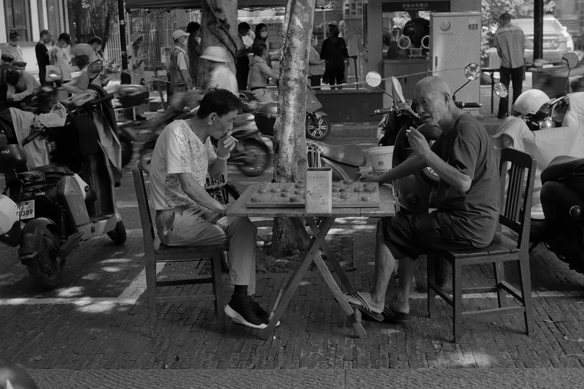 Two men sit across a small table playing chess inside a busy open-front workshop.