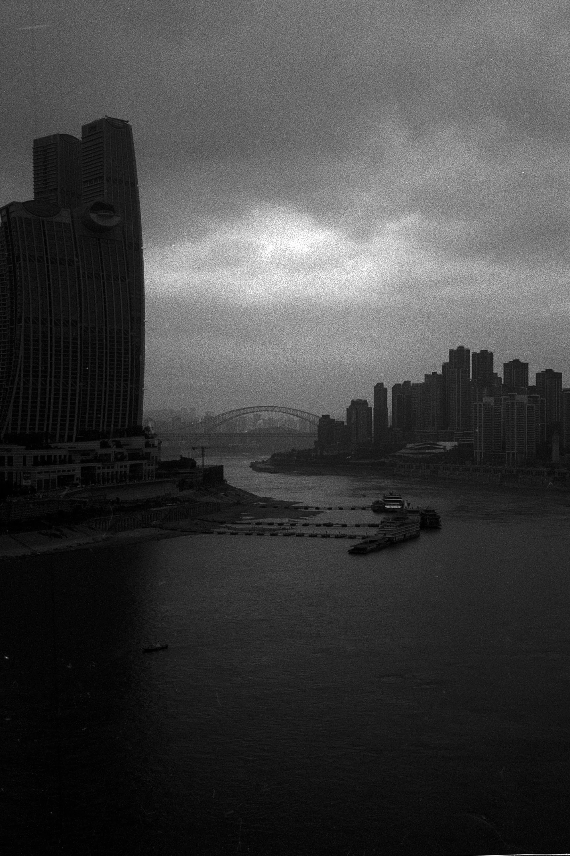 A tall dark building rises beside the river beneath dramatic storm clouds in Chongqing.