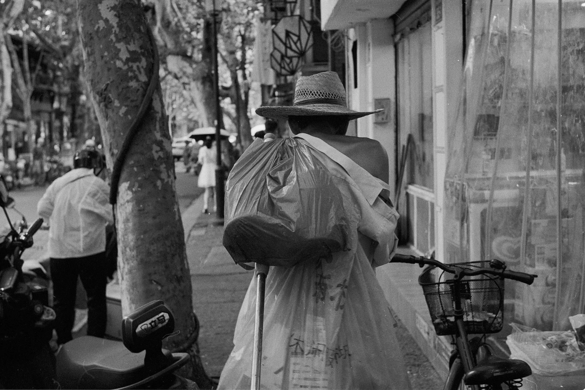A person in a brimmed hat walks along the pavement carrying several large bags.