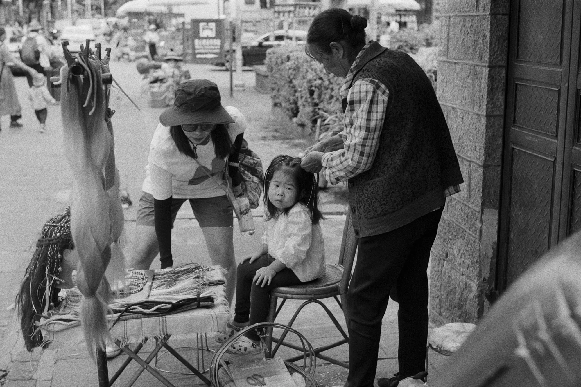 A child sits on a chair between adults and hanging goods at a busy market stall.