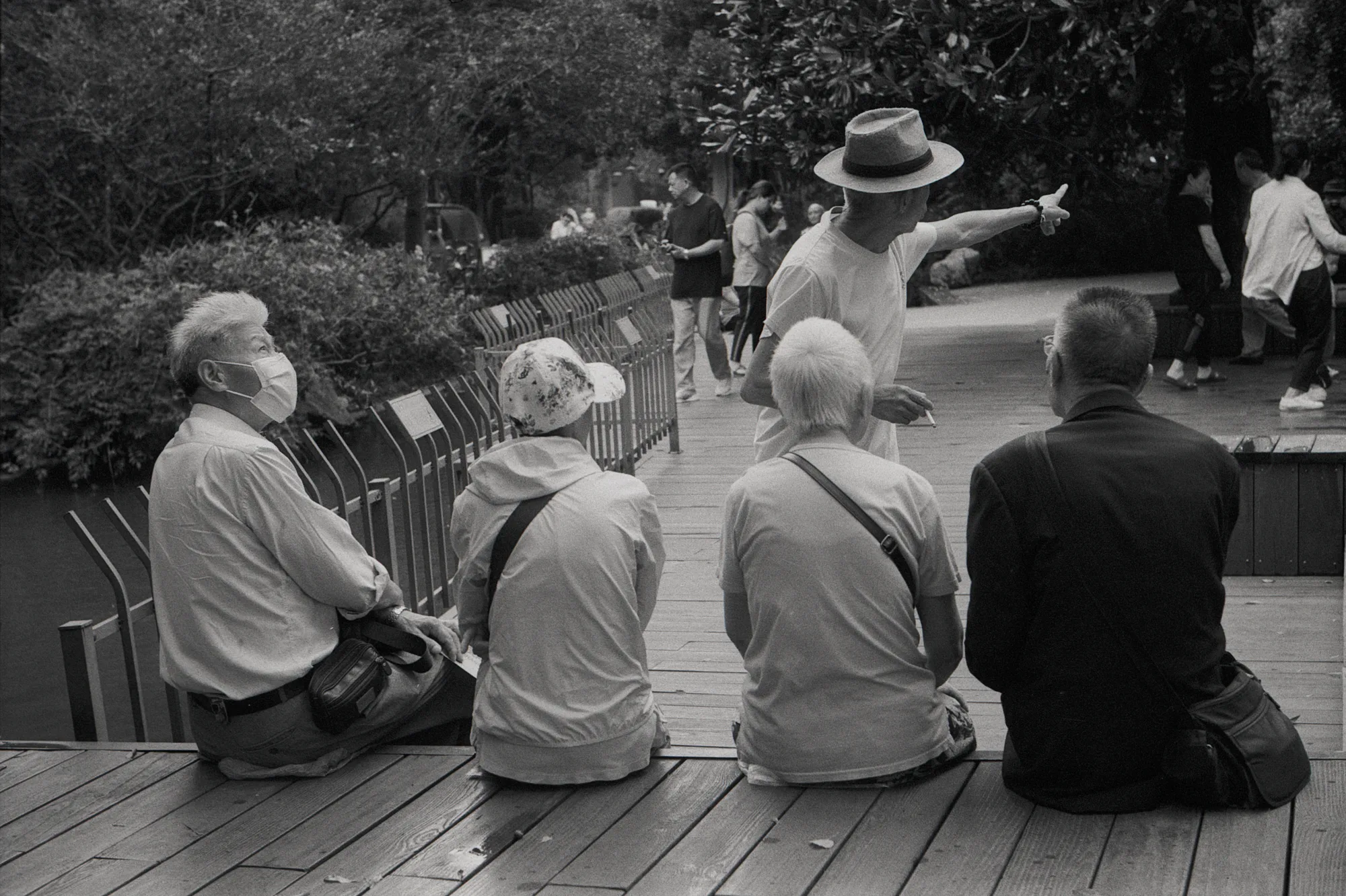 Four people sit on a wooden platform while a standing figure gestures near a fountain.