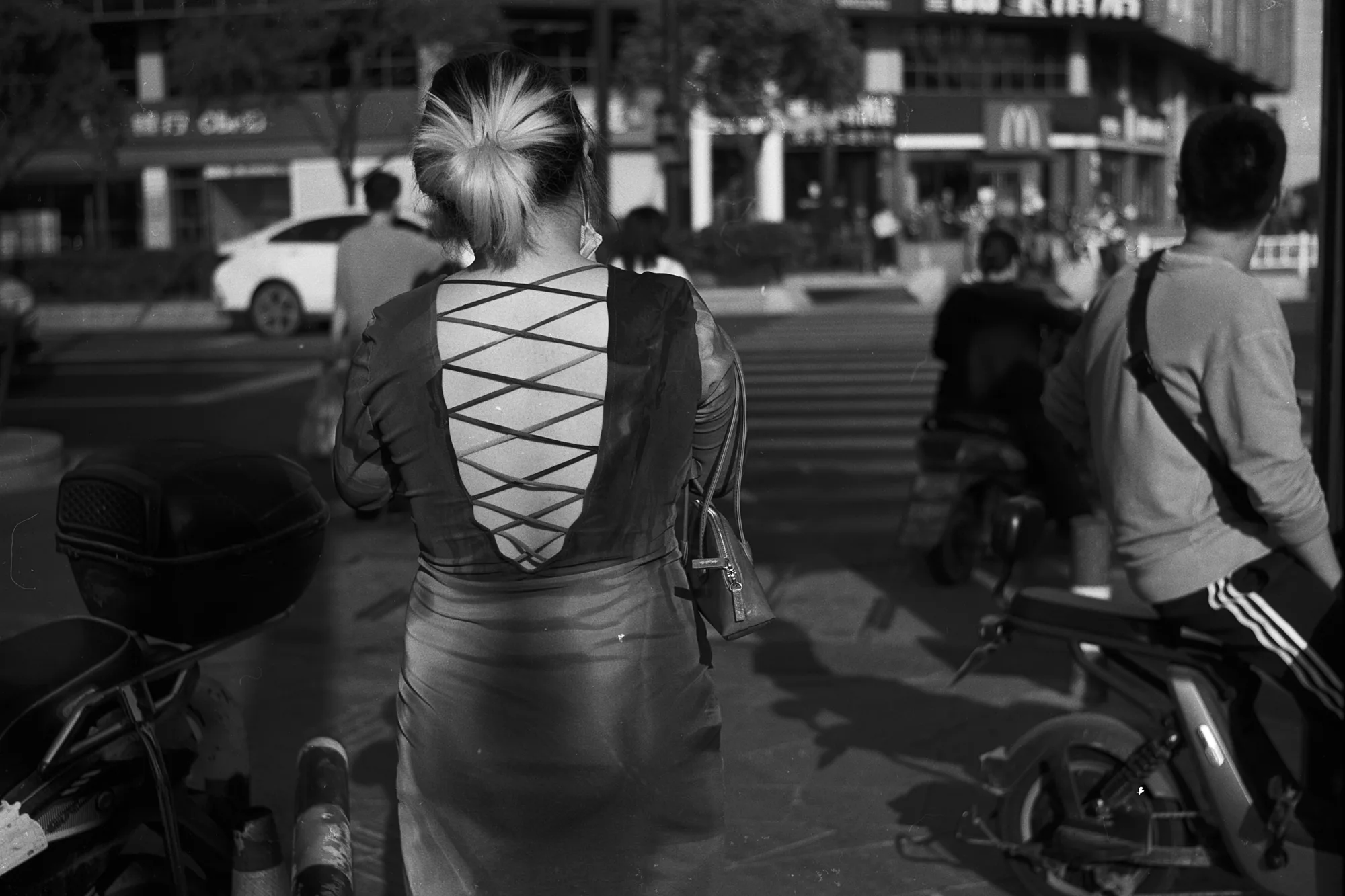 Cyclists wait at a street crossing, seen from behind a person wearing a laced-back top.