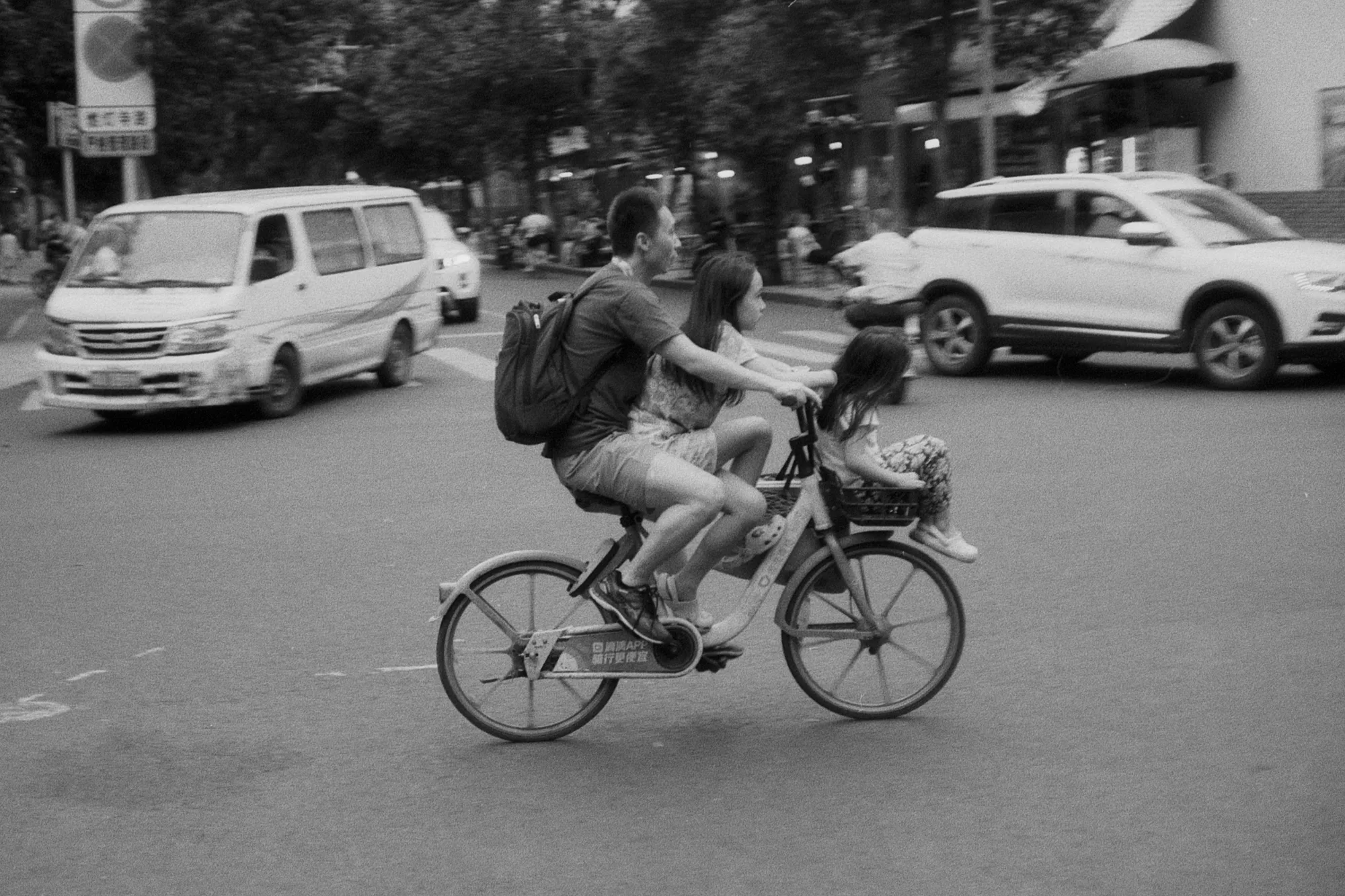 Two people ride together on a bicycle as cars and scooters pass around them.