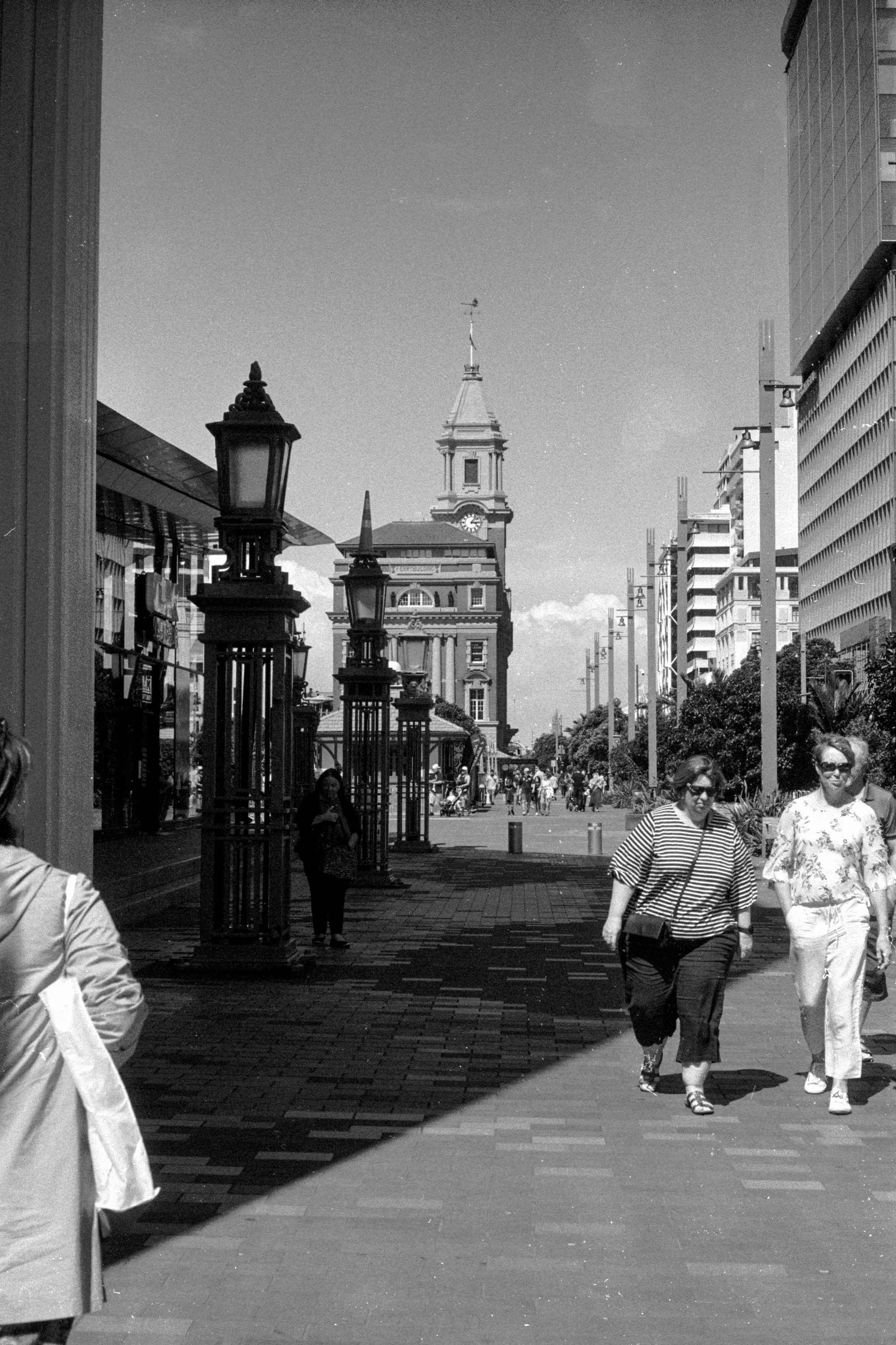 People walk through bright sunlight along Auckland's waterfront with city towers ahead.