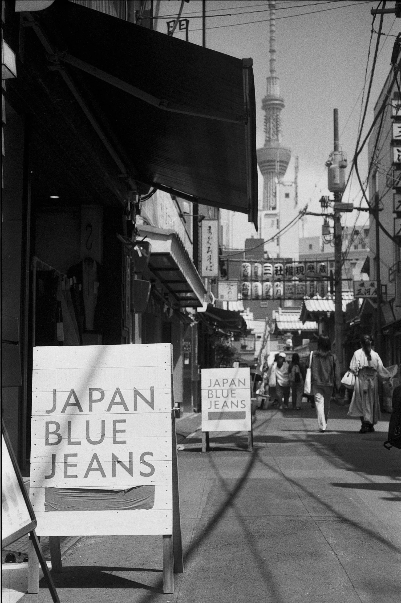 A sign reading Japan Blue Jeans stands at the entrance to a narrow sunlit lane.