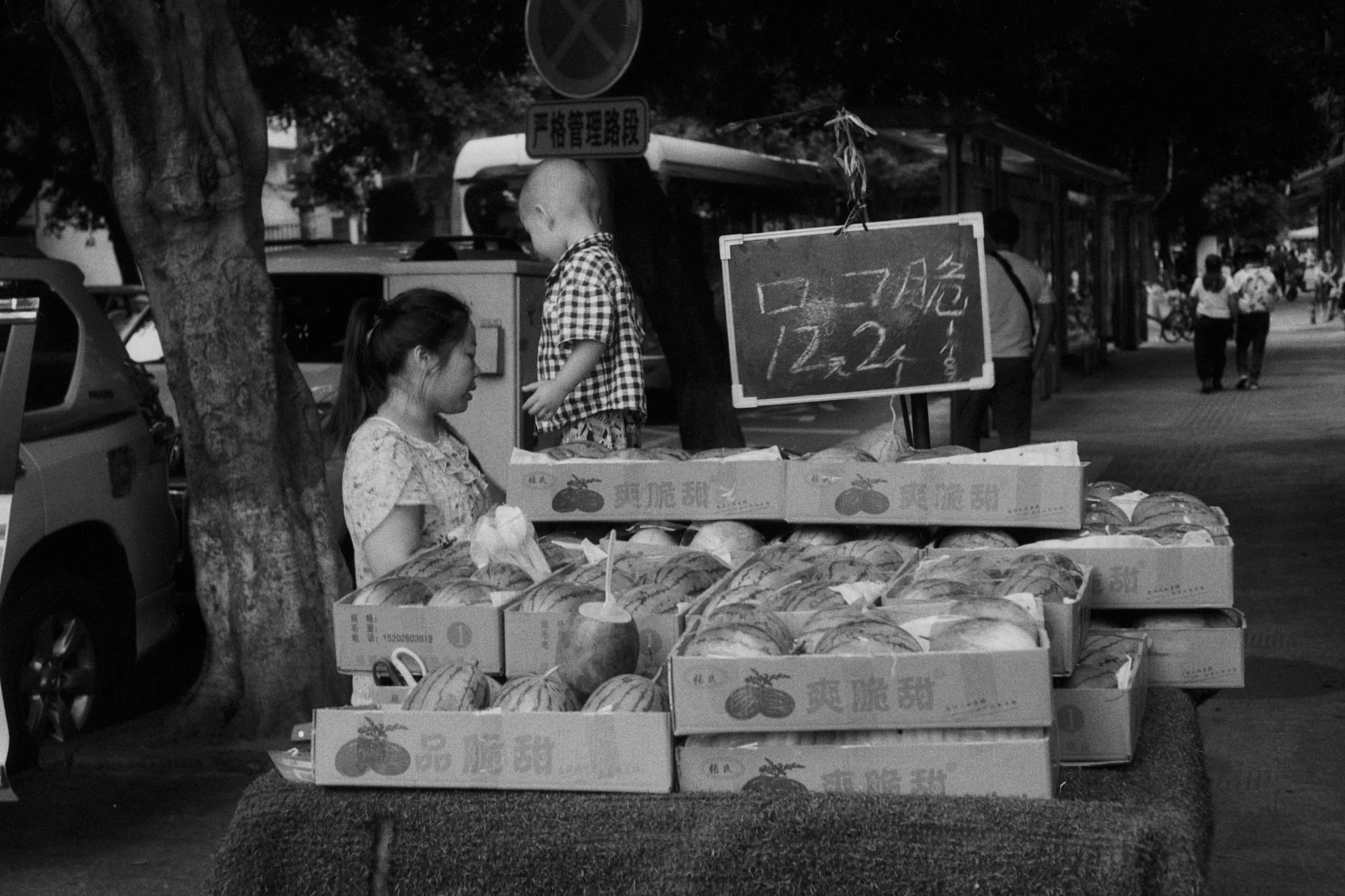 A seated vendor waits behind neat rows of fruit boxes at a Chengdu street stall.