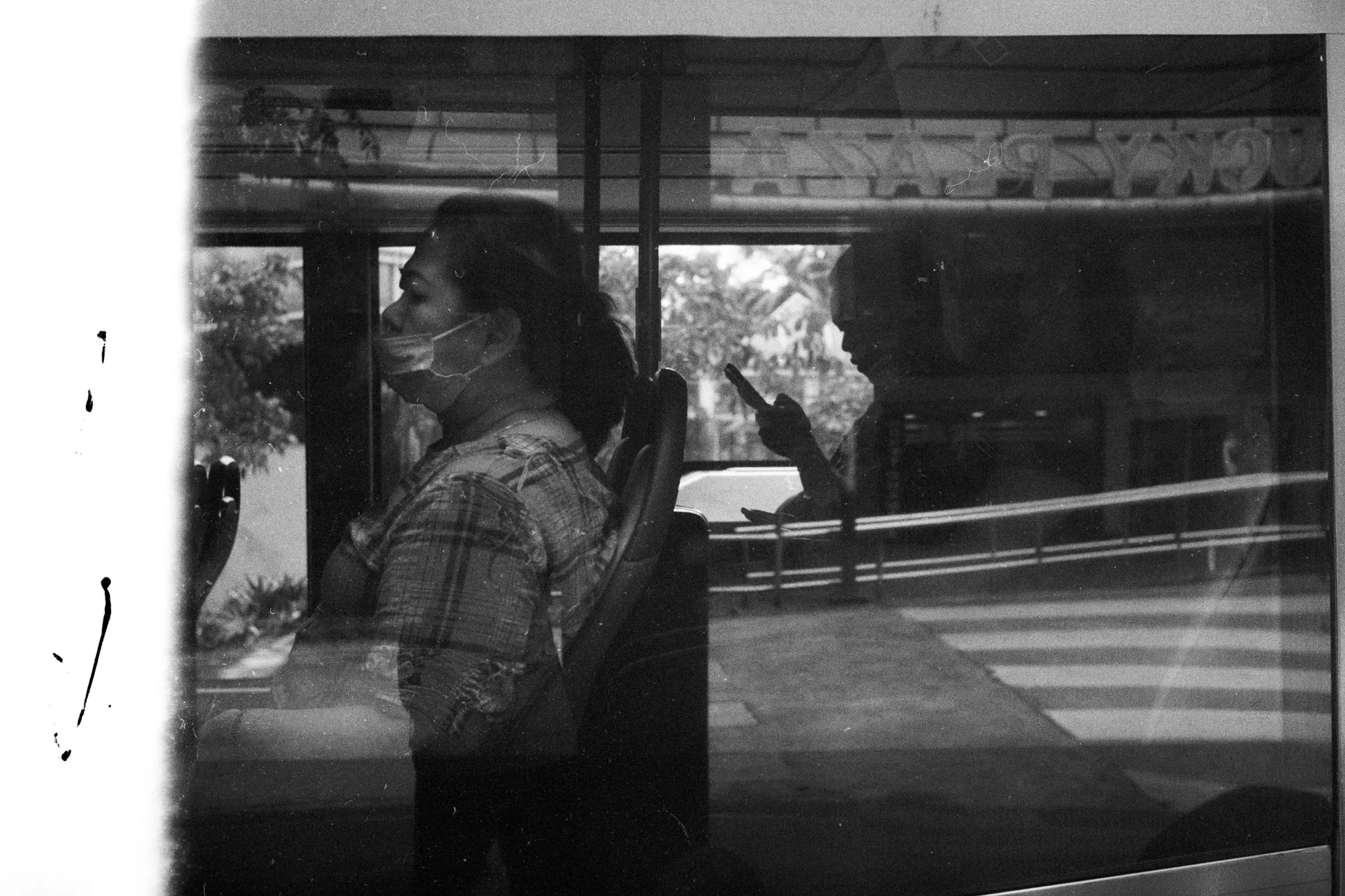 A passenger's profile overlaps with trees and street reflections in a bus window.
