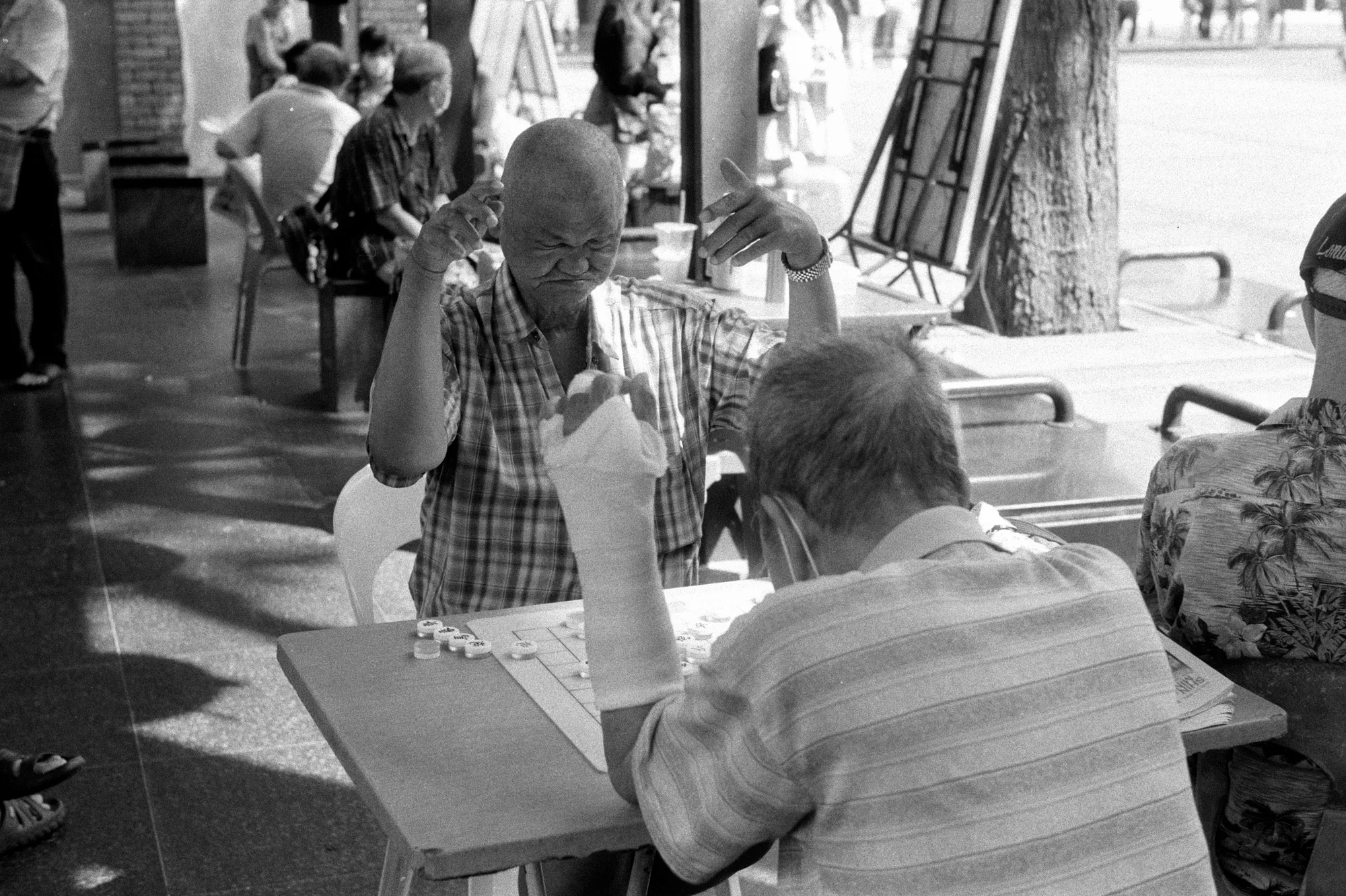 Two older men sit across a small table playing chess beneath the shade of street trees.
