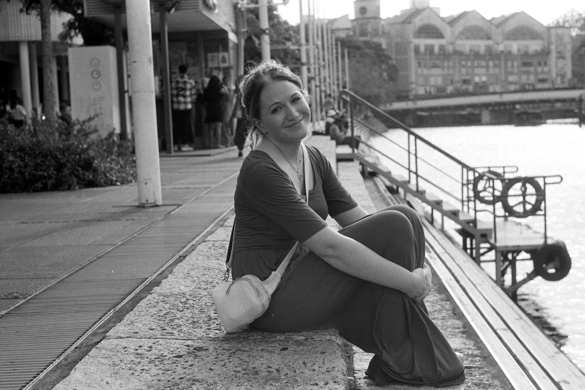 A woman sits on a low ledge beside the water at Clarke Quay and looks toward the camera.