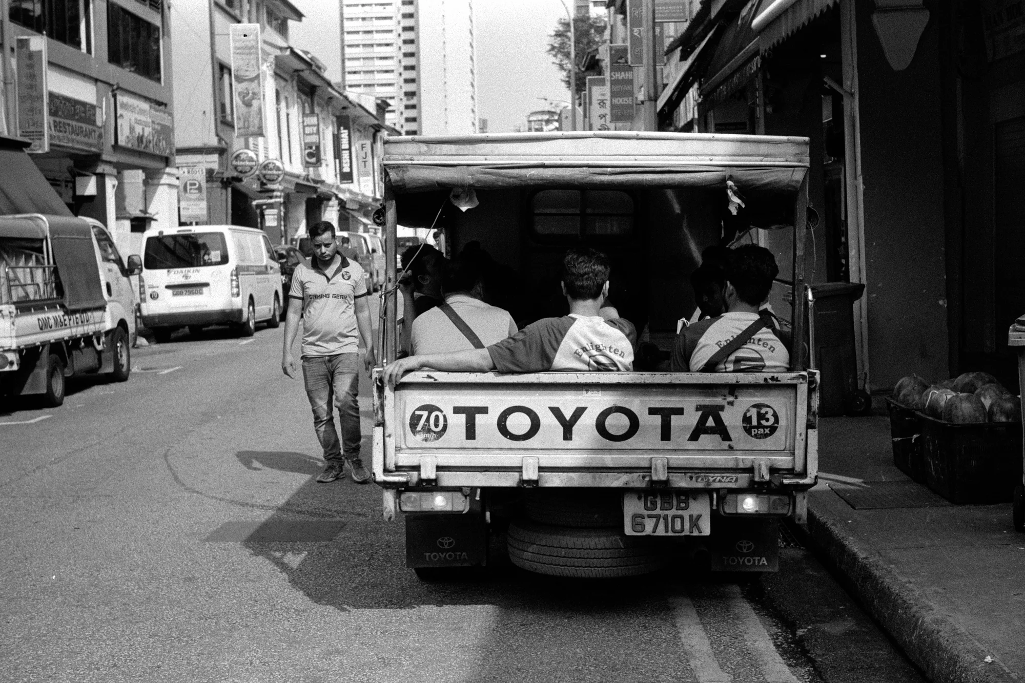 A small Toyota truck drives along a narrow lane lined with shops and parked vehicles.