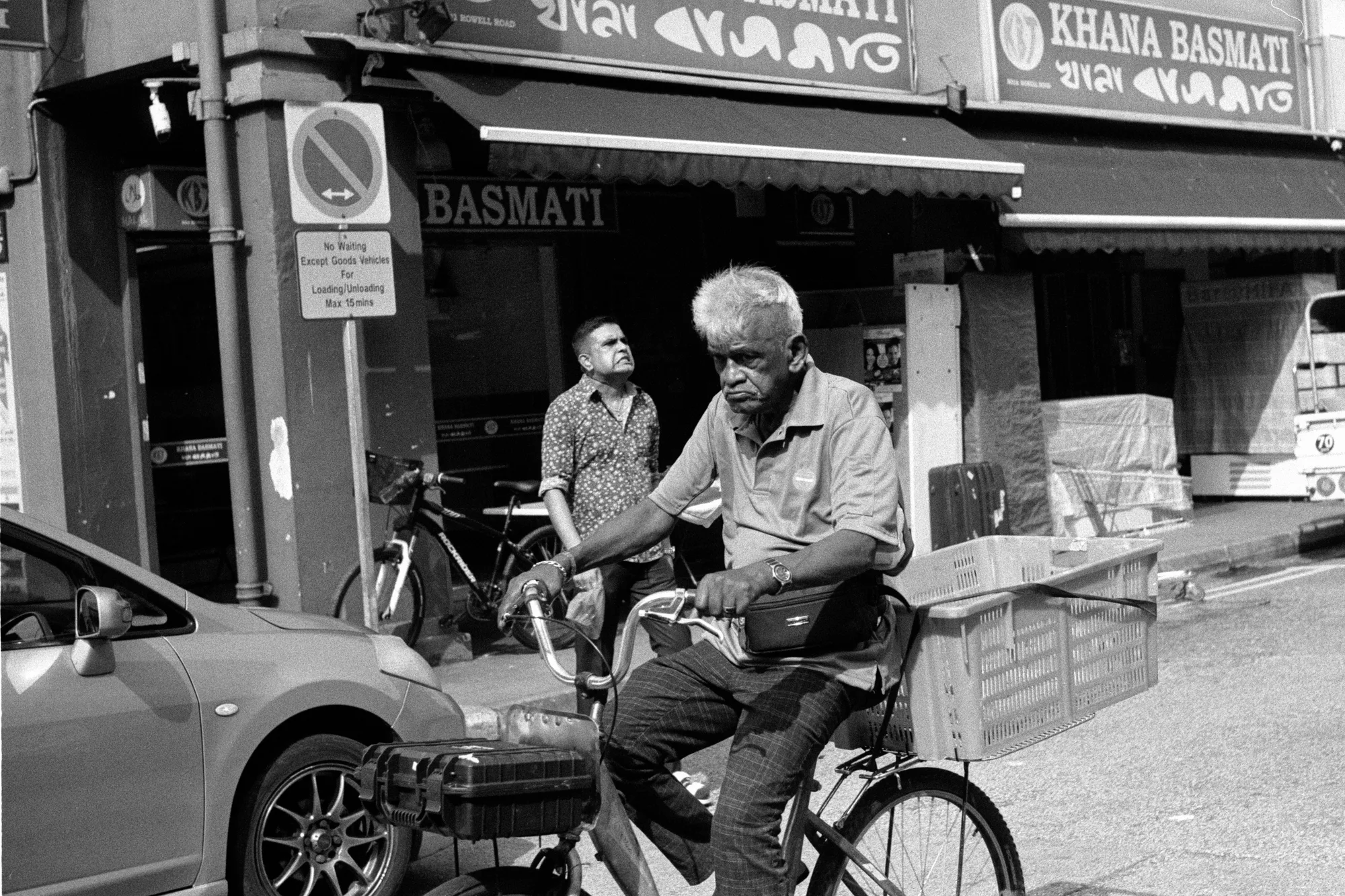 An older cyclist rides past a corner shop while two pedestrians stand behind him.