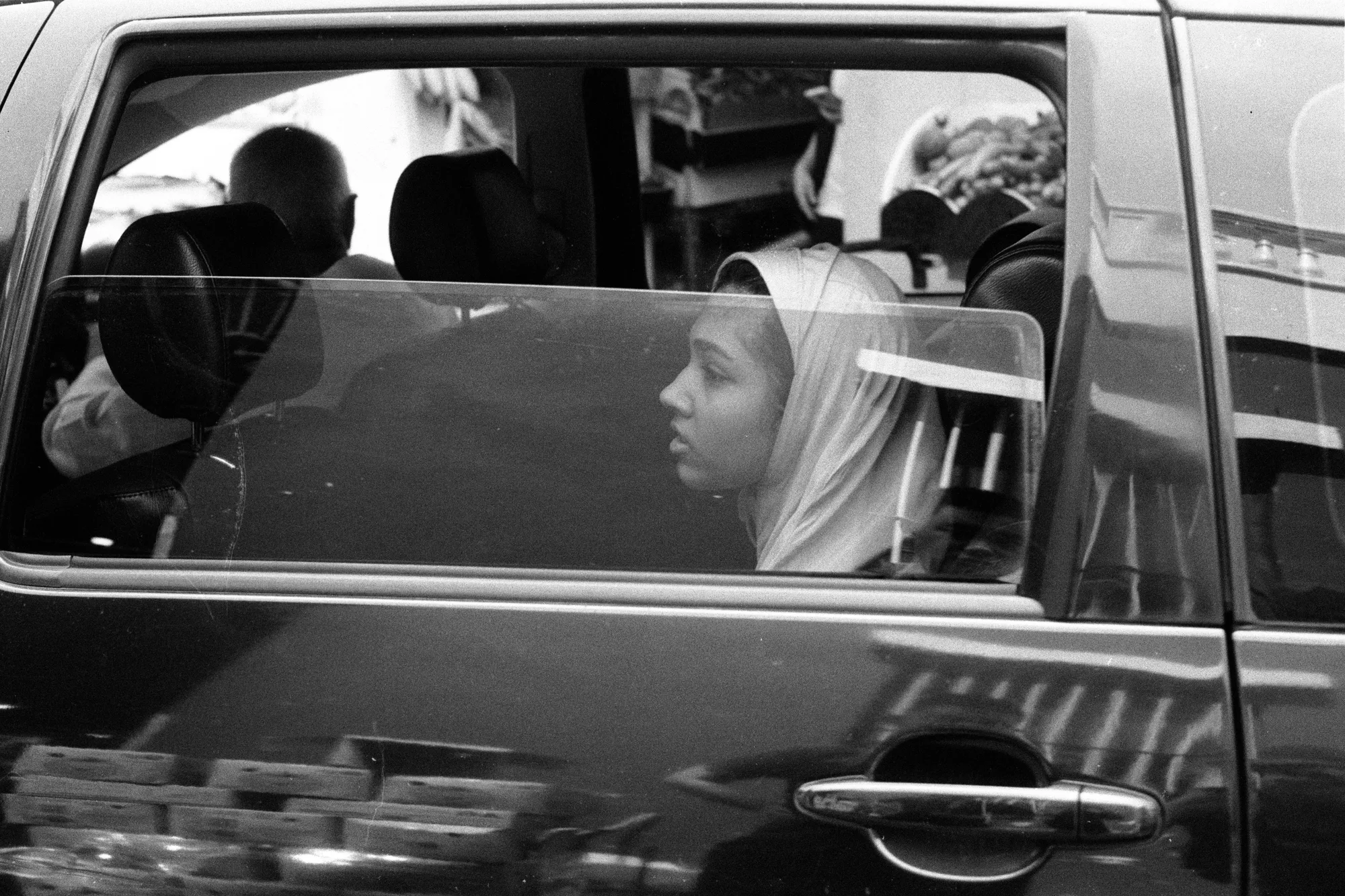 A young passenger wearing a headscarf looks through the open rear window of a car.