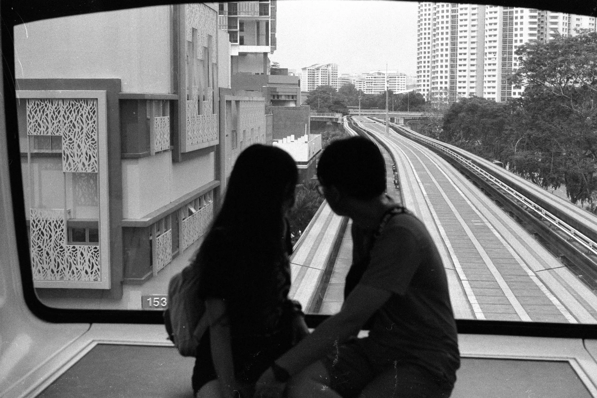 Two young passengers lean close together beside a large window overlooking the rail tracks.