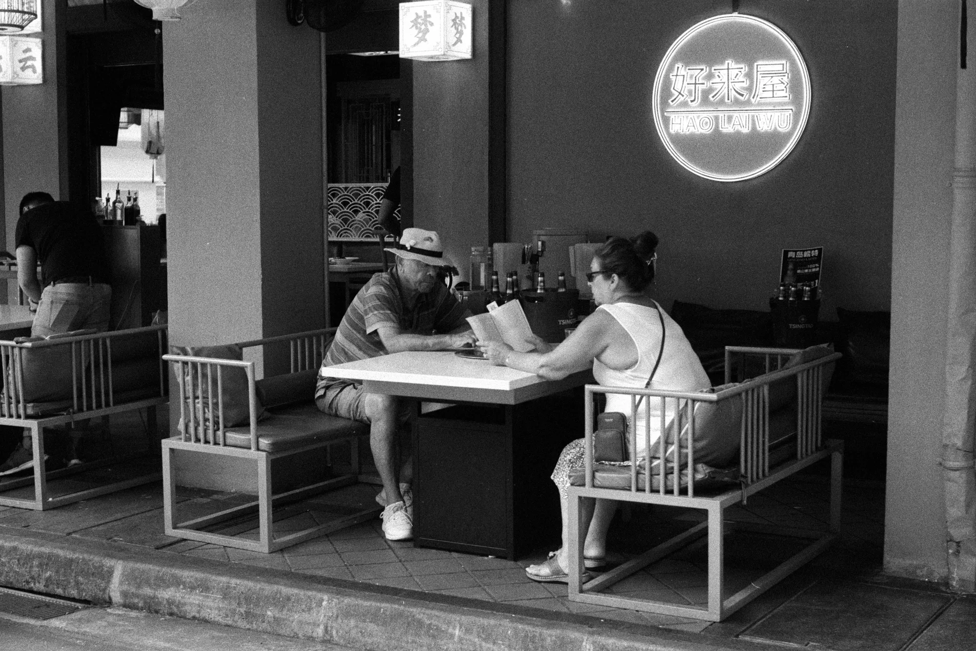 Two people sit across from each other reading at a table inside an open-front cafe.