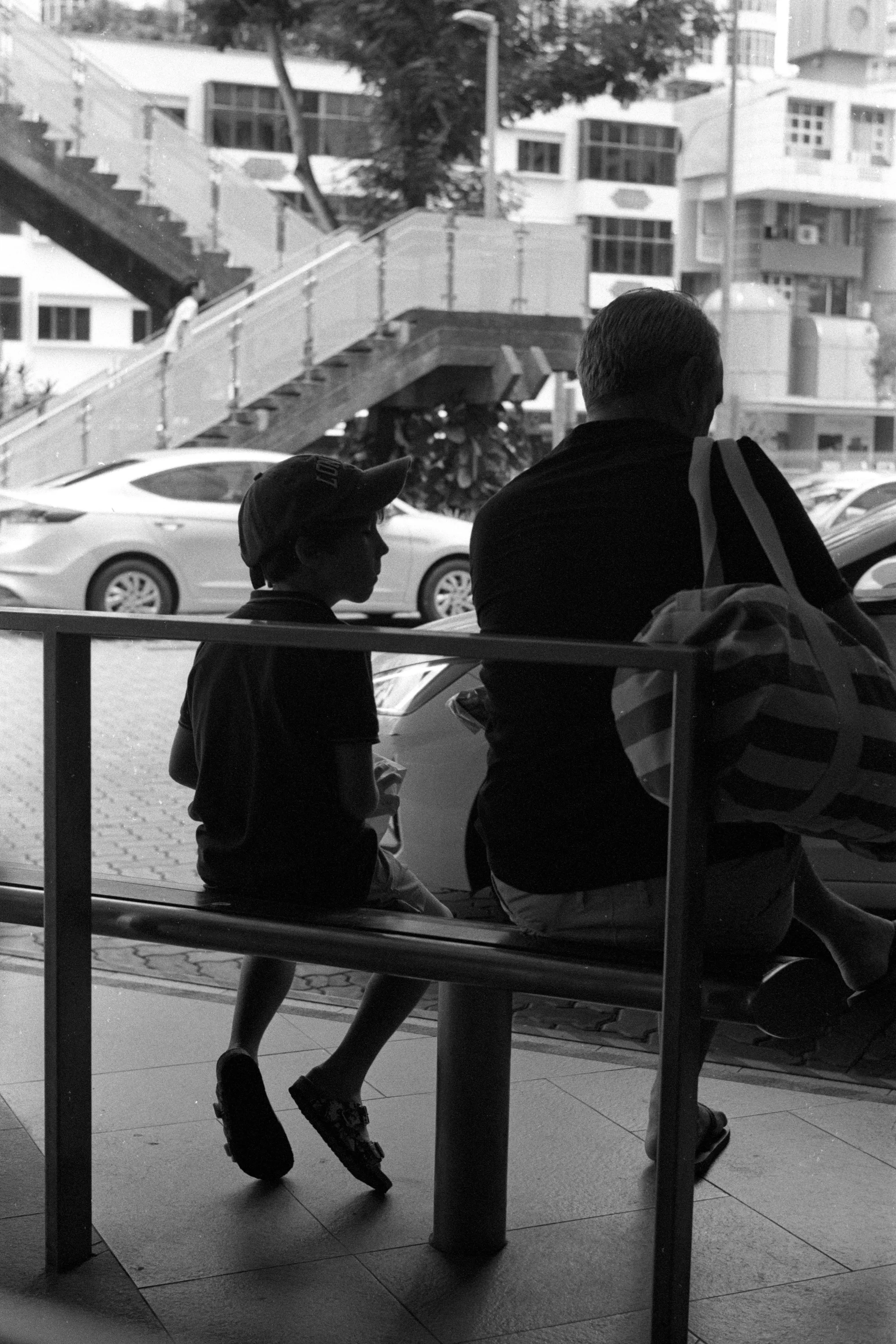 A child stands beside a seated adult at a bus stop, seen through a window.