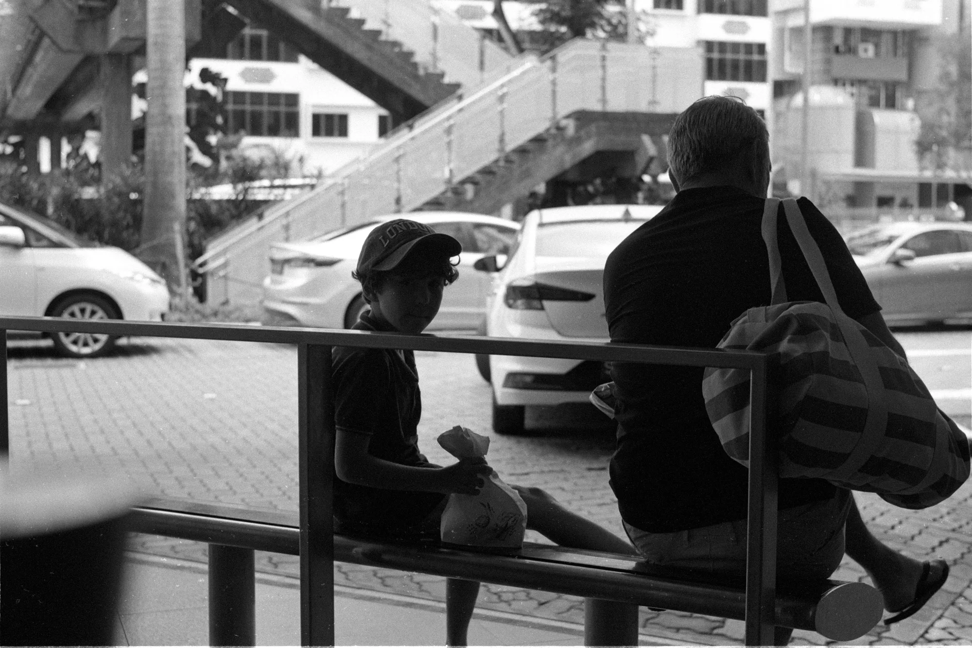 A child crouches near a seated adult while a white car passes behind them.