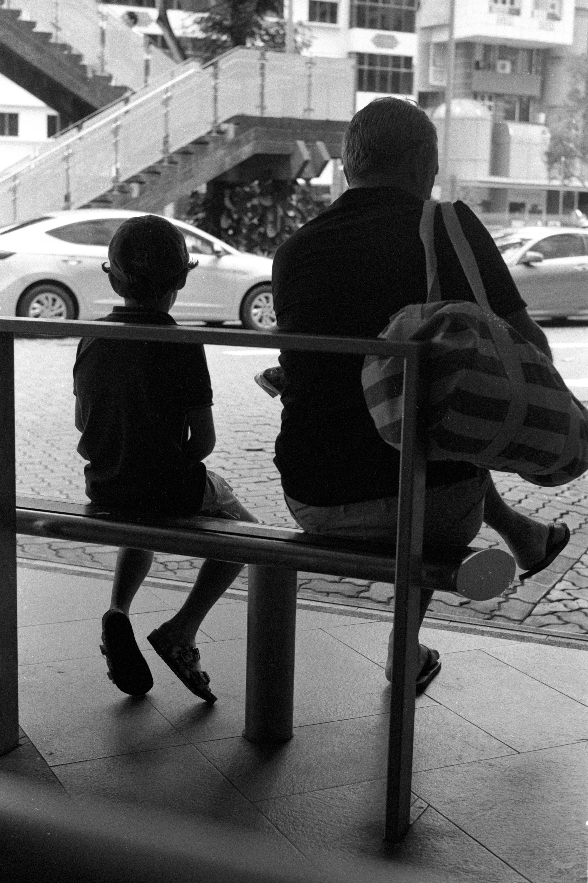 A child stands close to a seated adult behind the bus stop railing.
