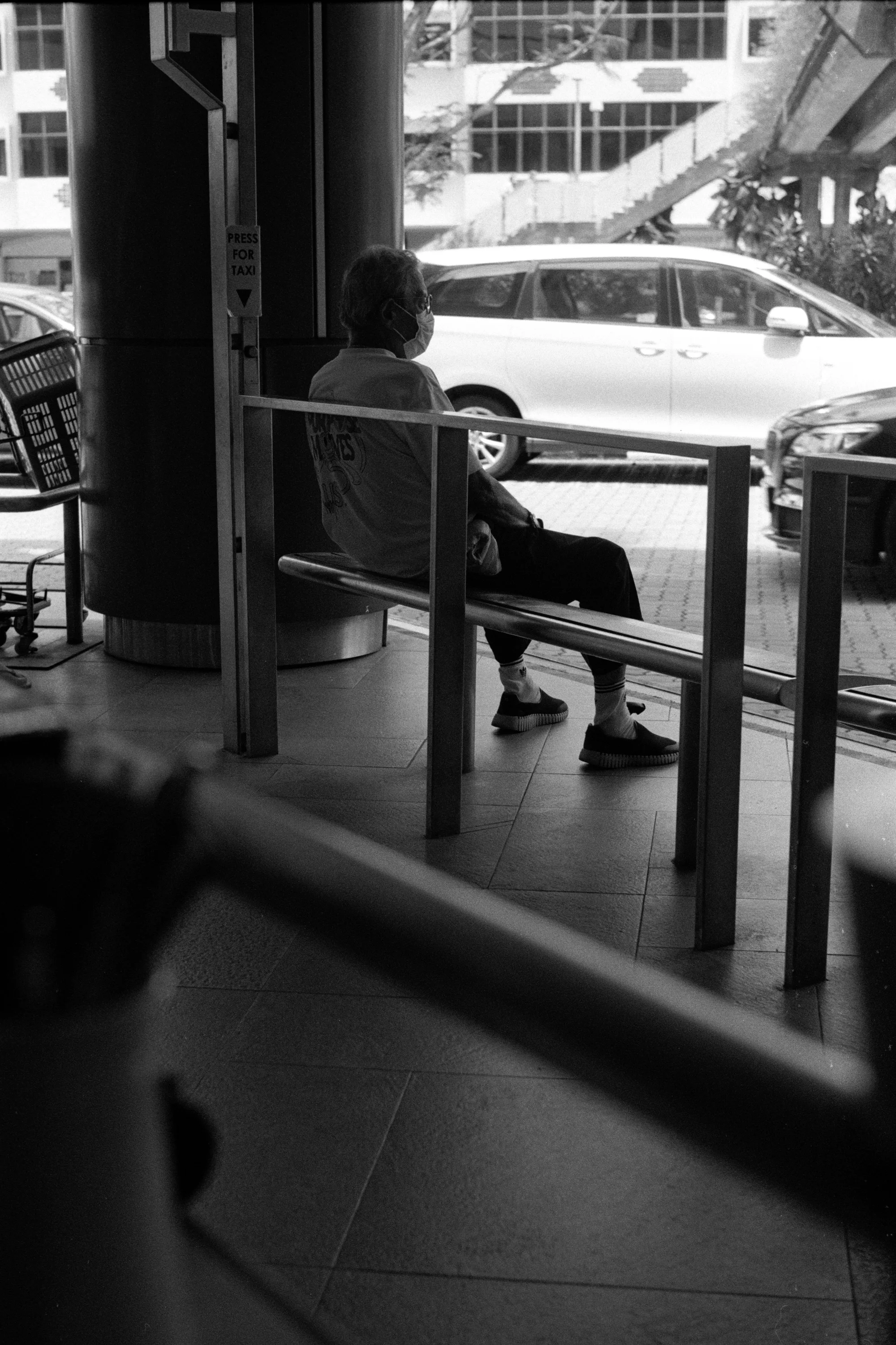 A child is partly hidden by a railing beside a seated adult at the bus stop.
