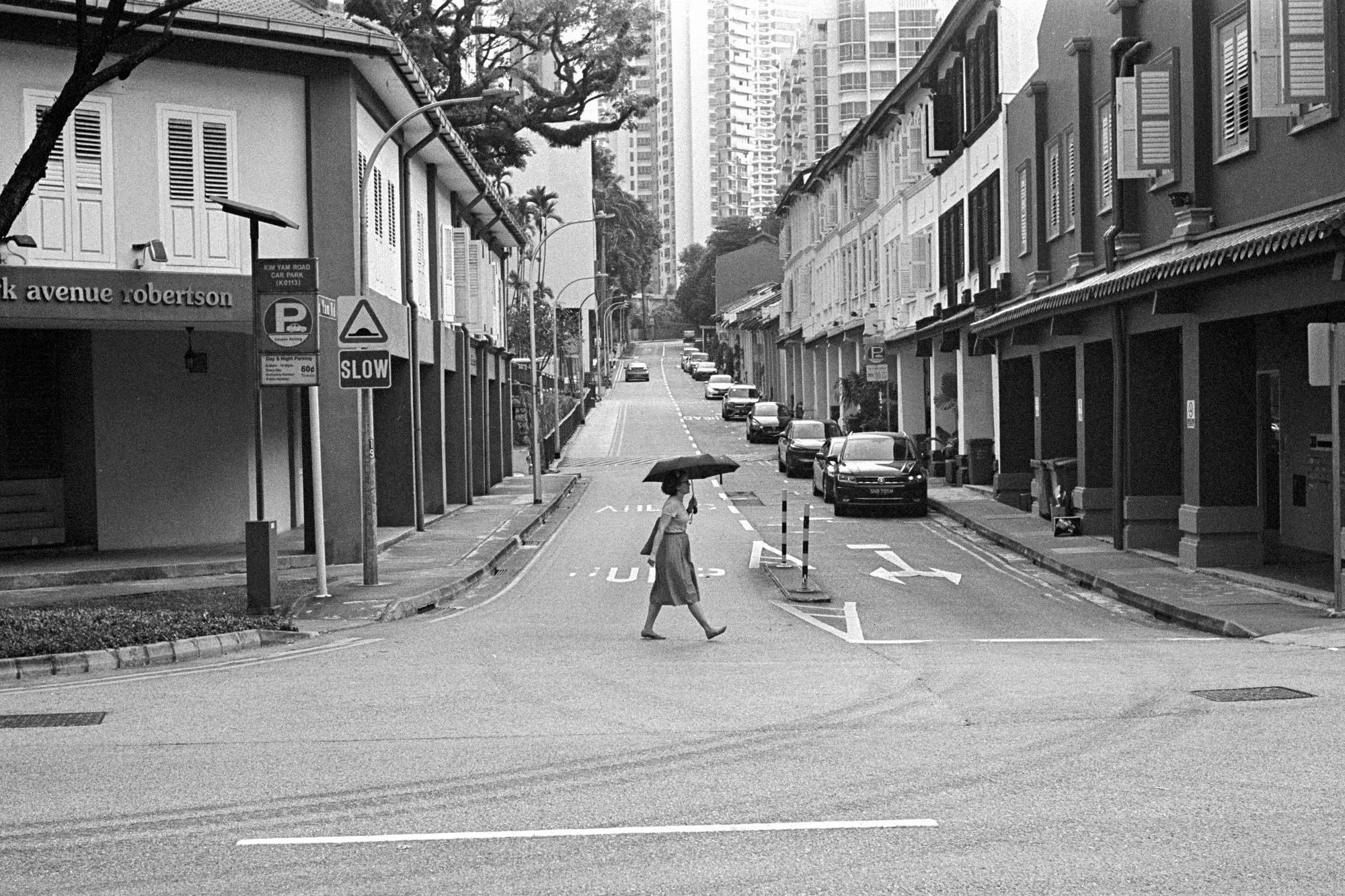 A lone pedestrian crosses a quiet street between low buildings and parked cars.