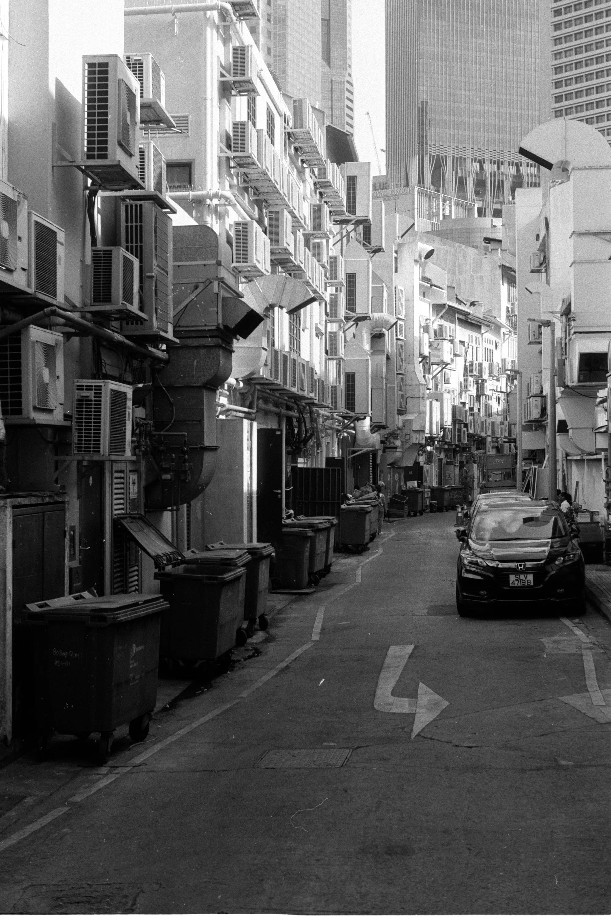 A narrow street lined with air conditioners and service entrances leads toward city towers.