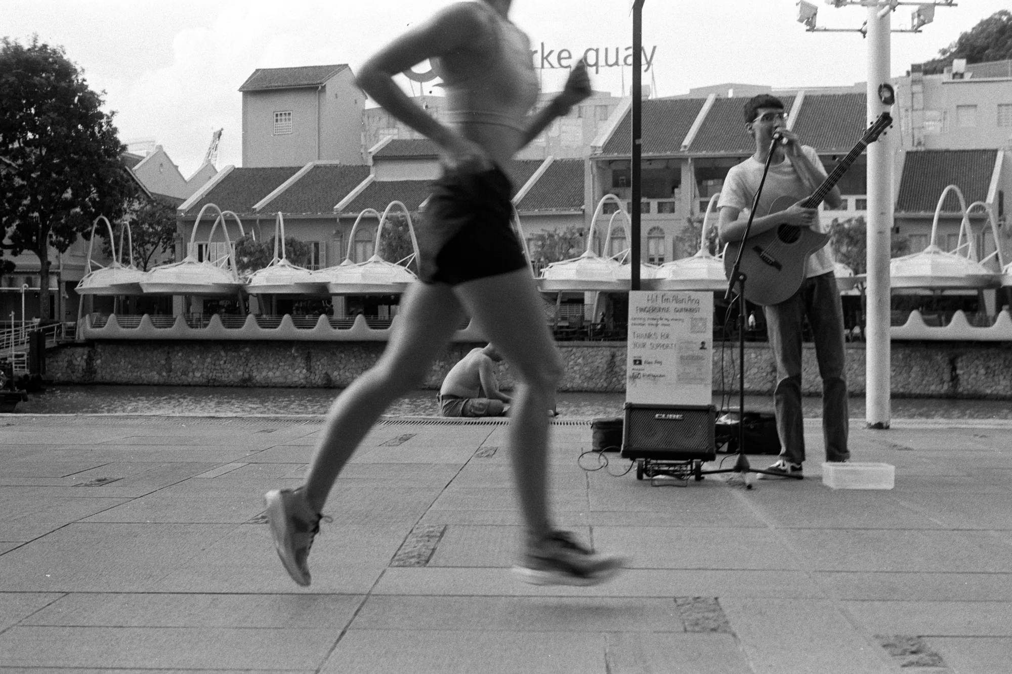 A blurred runner crosses in front of a street musician performing beside the river.