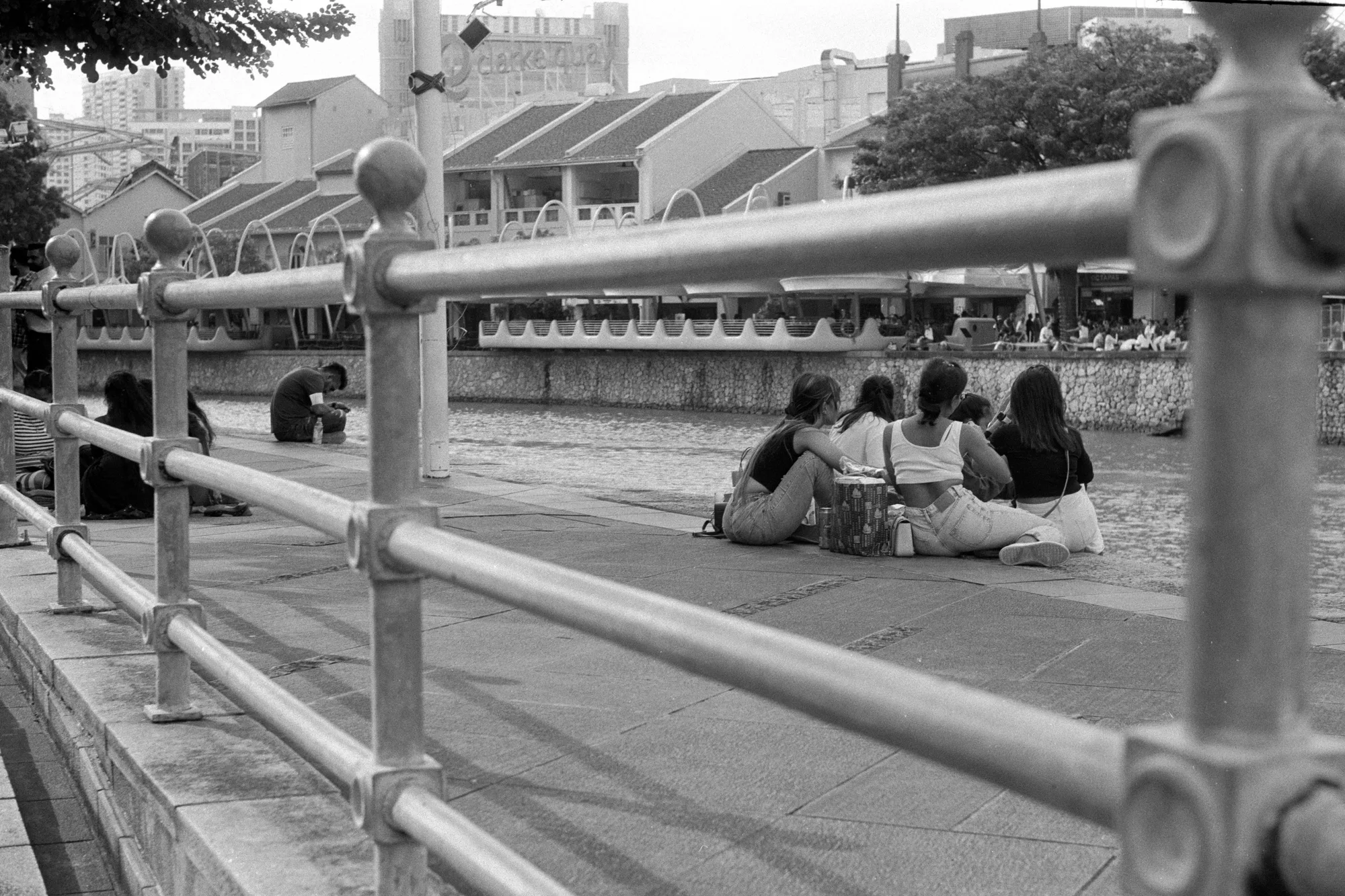Four people sit together on the pavement beside a riverside railing.