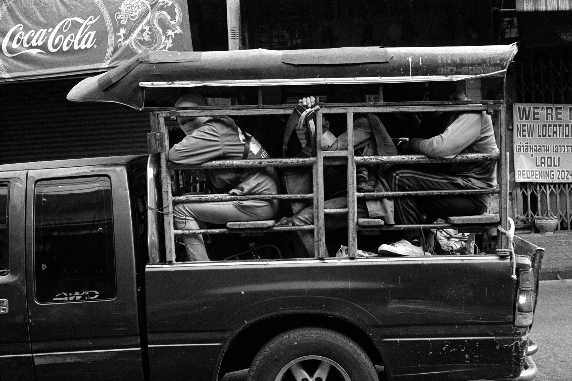 Several passengers sit behind metal rails in the open back of a truck.