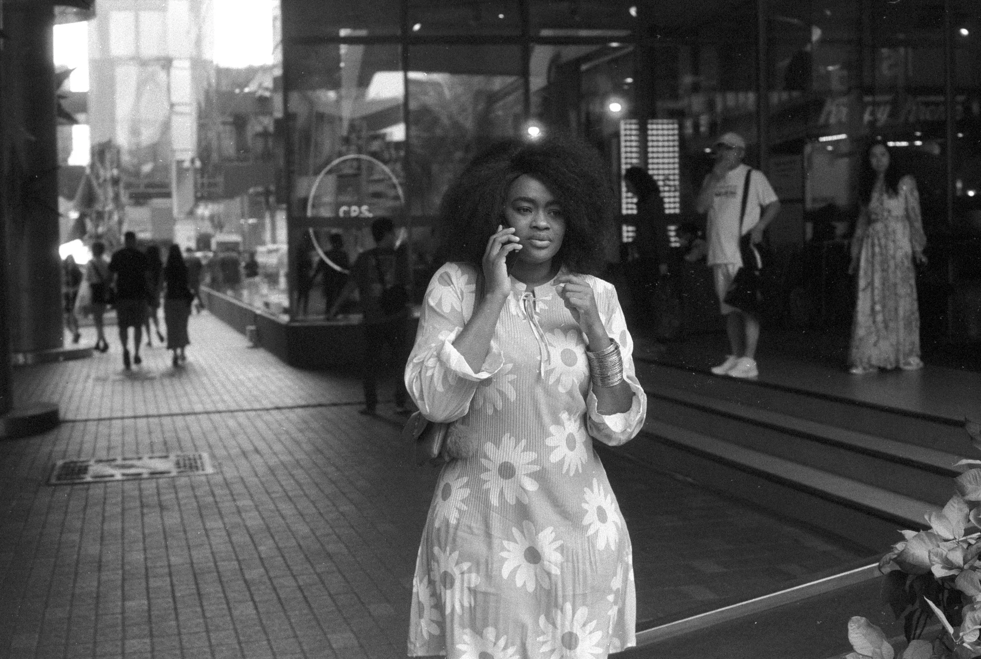A woman in a patterned dress looks at her phone on the pavement outside a shop.
