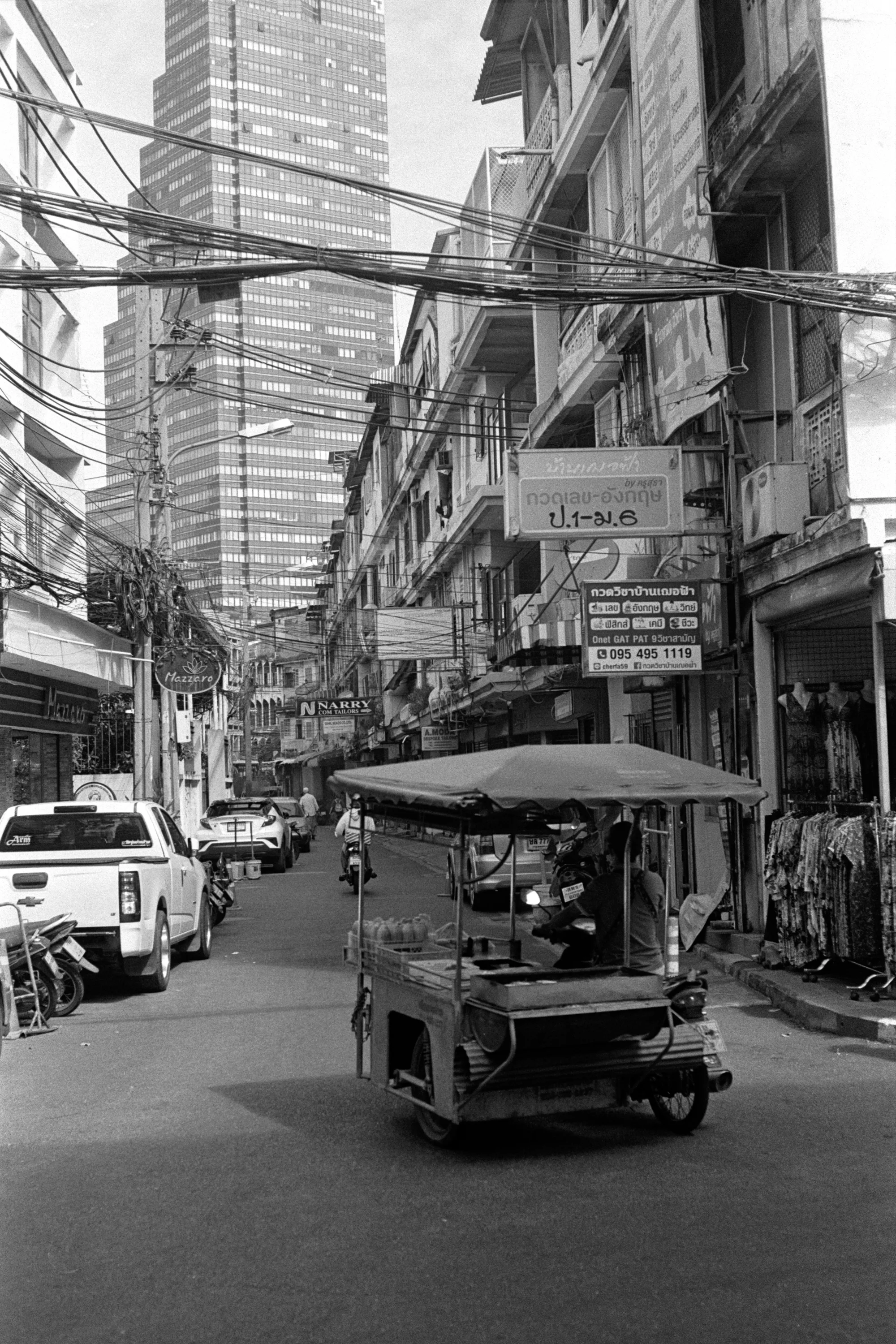 A tuk-tuk drives down a narrow street beneath dense overhead utility wires.