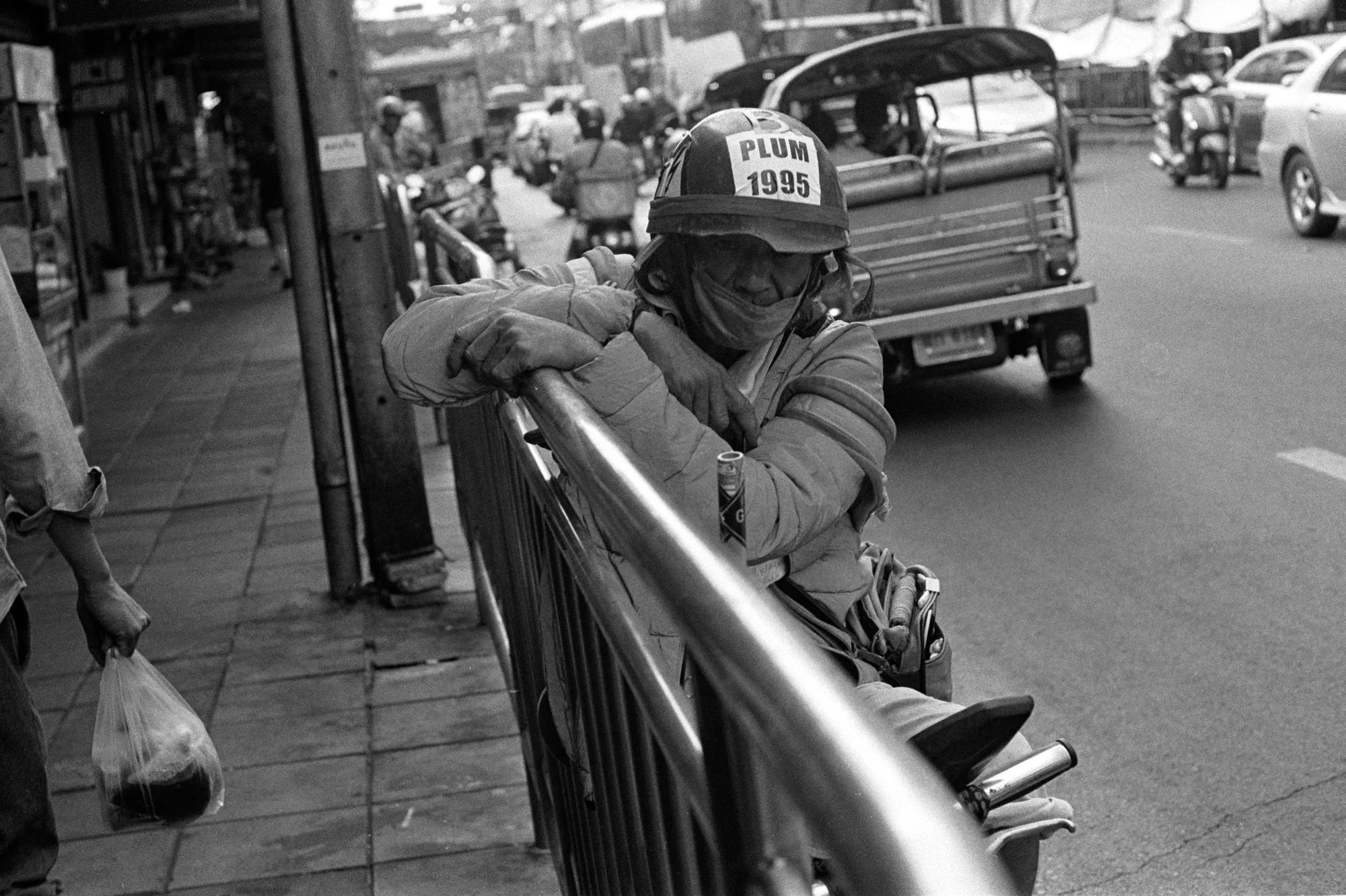 A man in a cap leans on a roadside rail with tuk-tuks and traffic behind him.