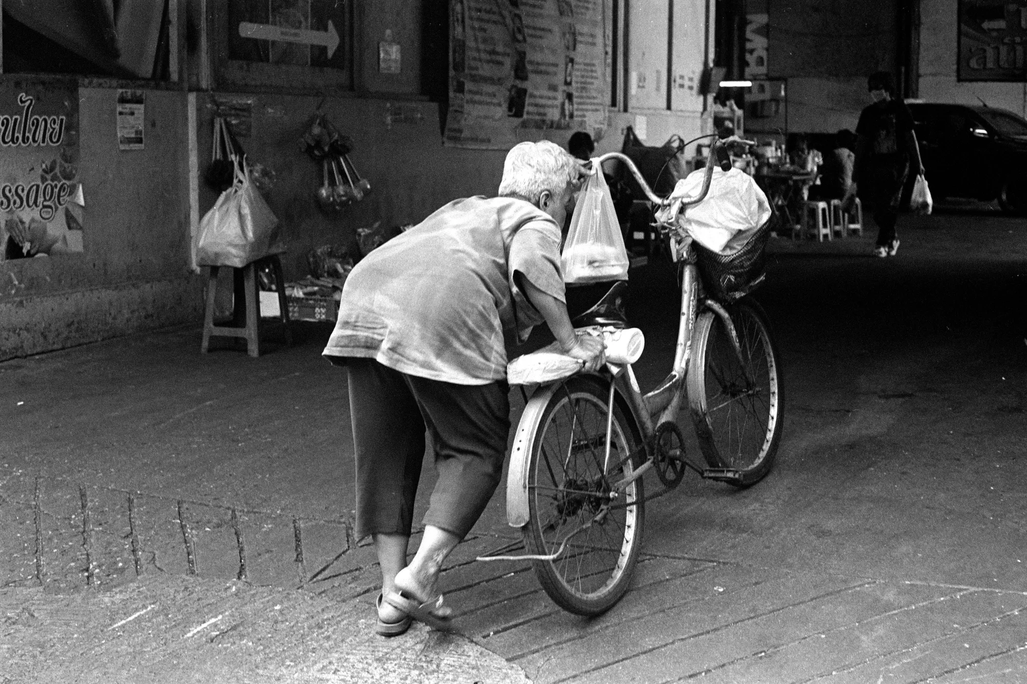 A person bends over a bicycle while another bicycle stands nearby on a side street.