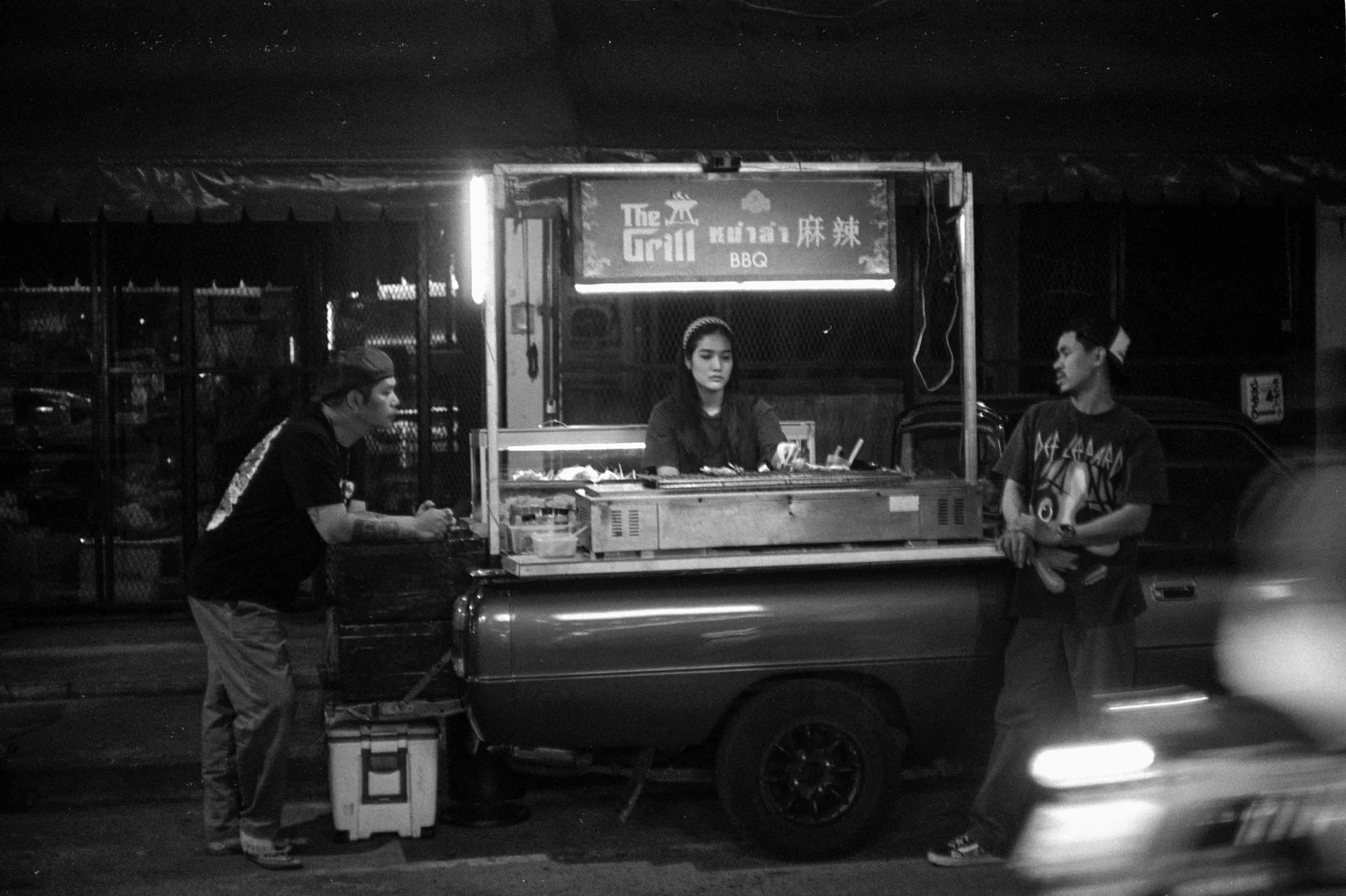 A vendor stands inside a lit food cart while customers wait beside it at night.