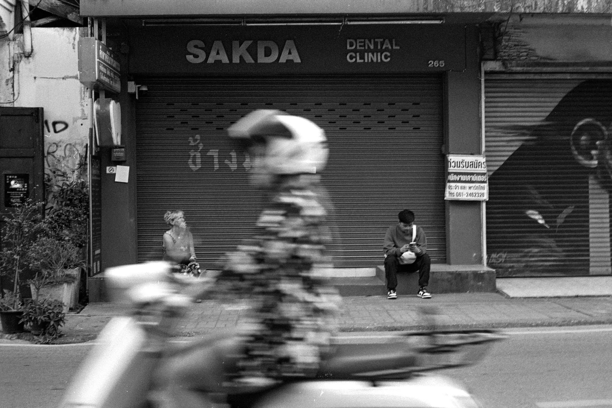 A blurred motorbike passes two children seated outside a closed storefront.