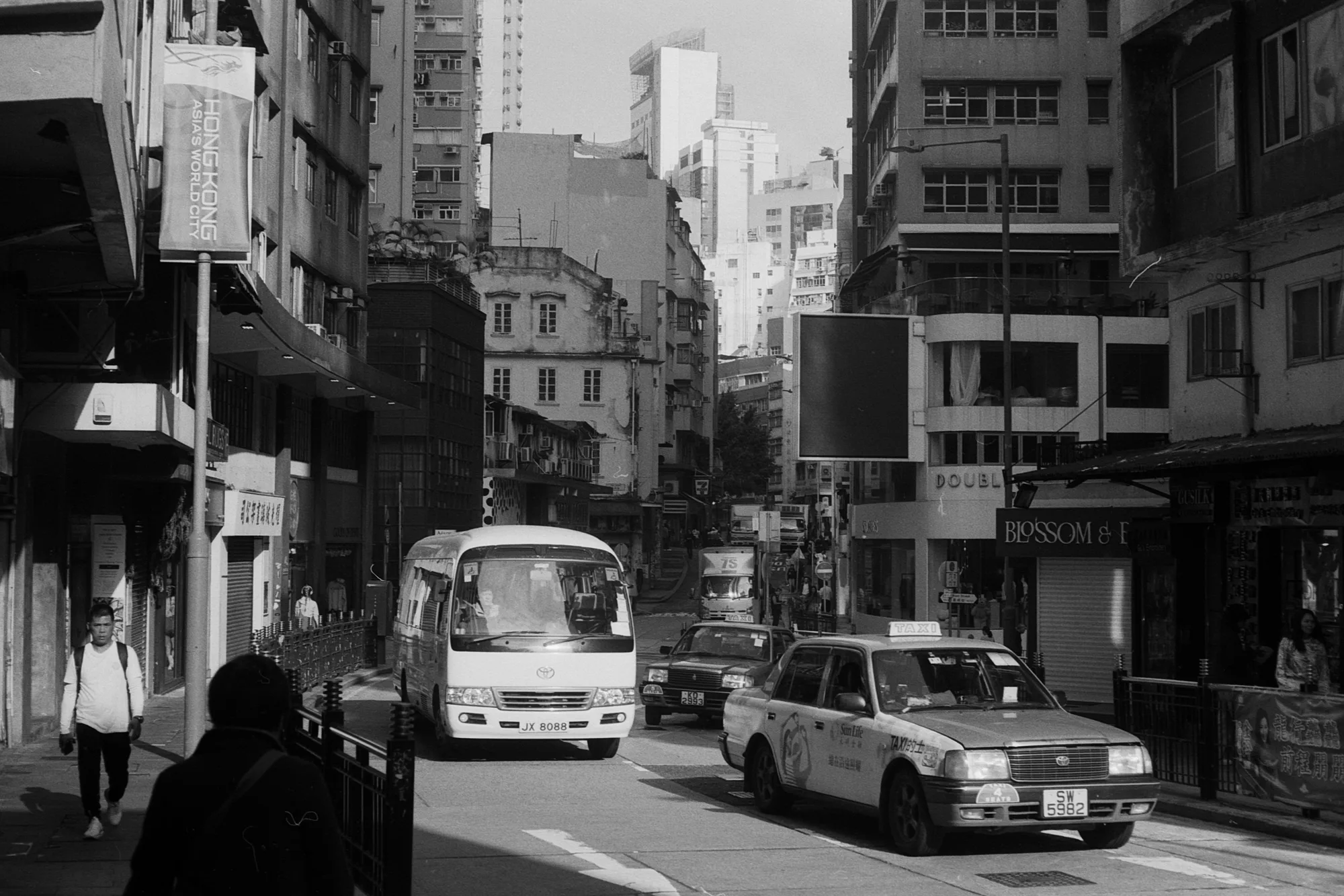 Cars and a minibus travel along a Hong Kong street lined with older buildings and towers.