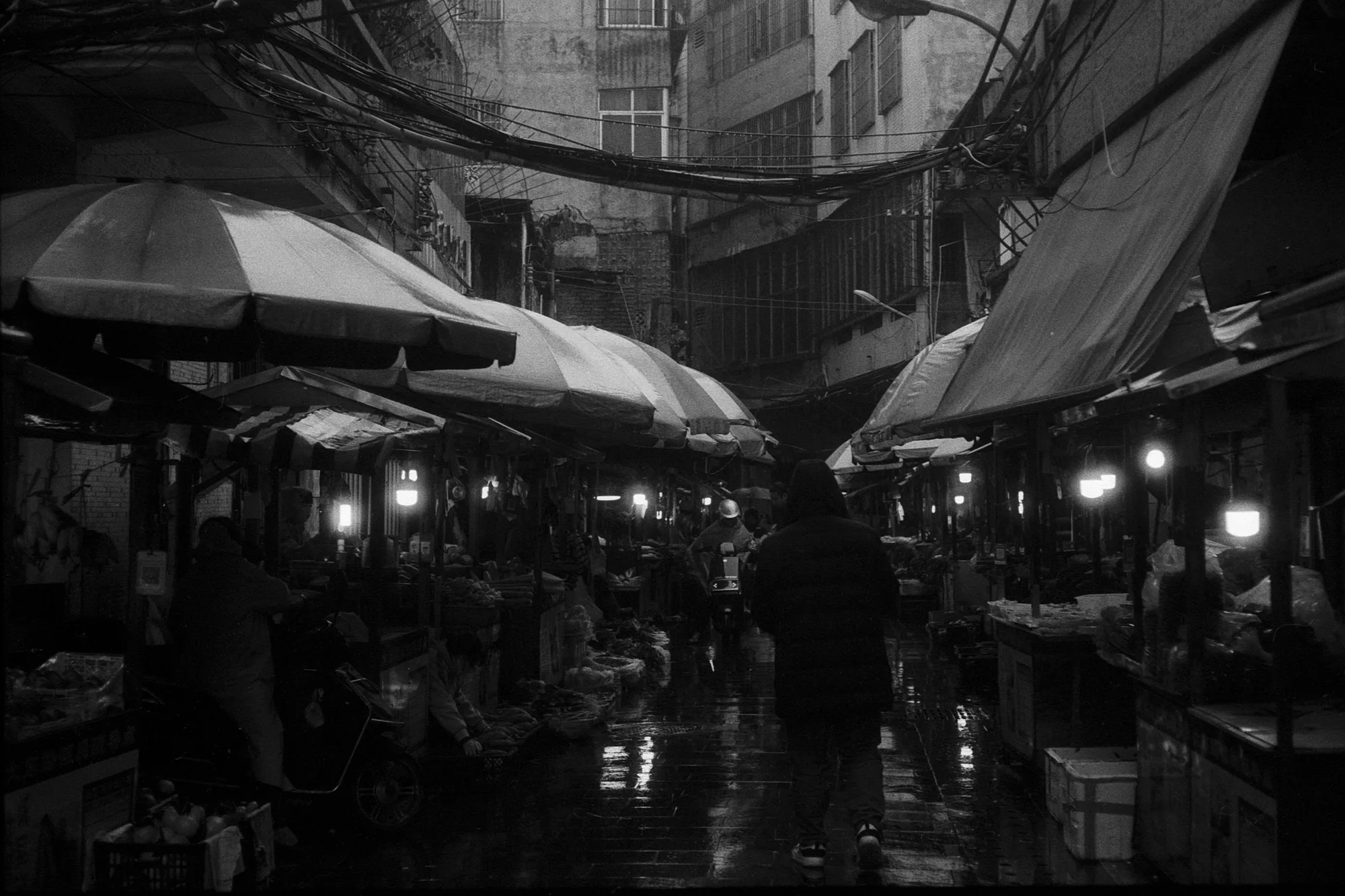 Lighted market stalls sit beneath umbrellas along a narrow street at dusk.