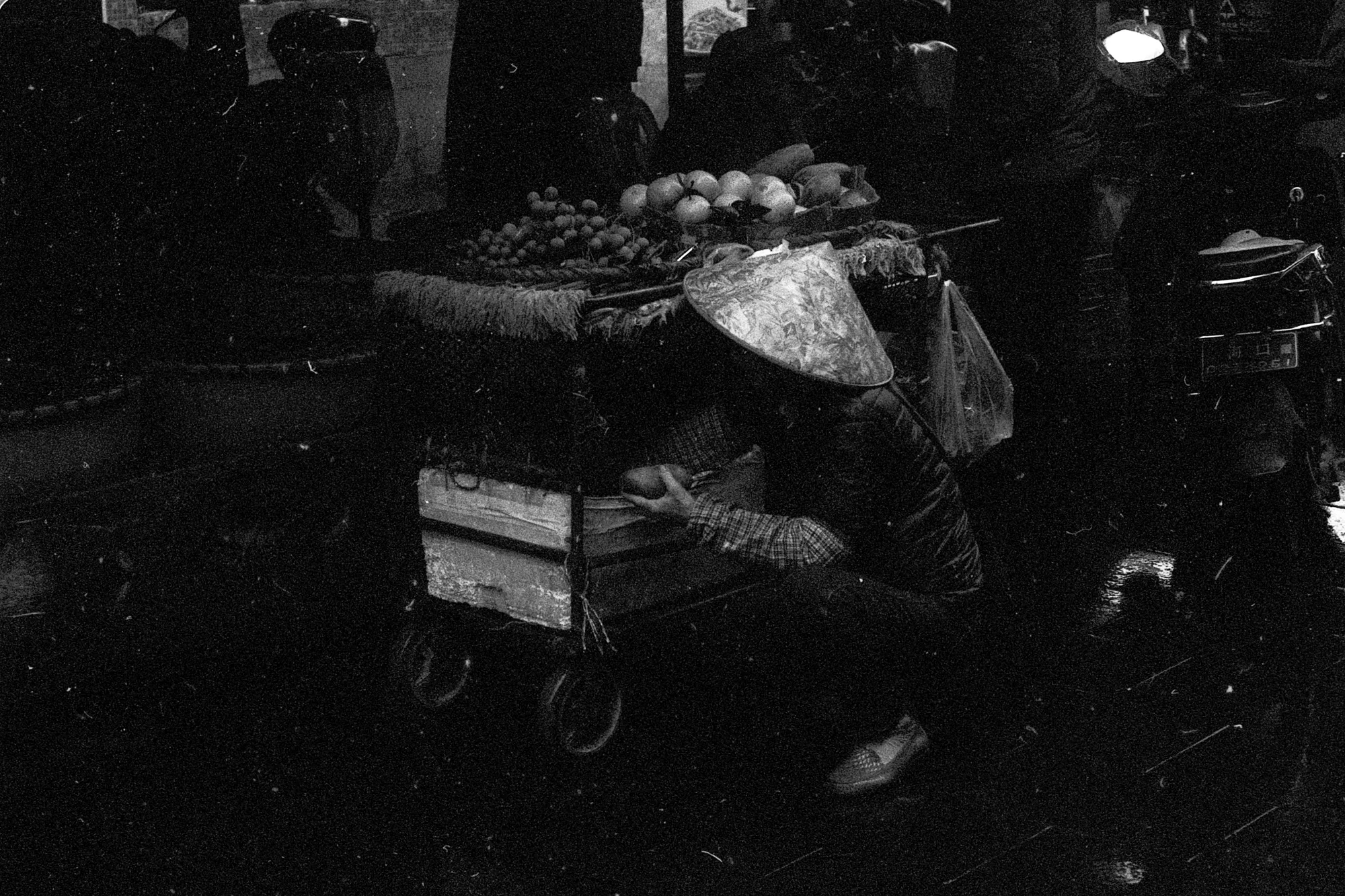 A vendor stands behind a produce cart in the deep shadows of a market.