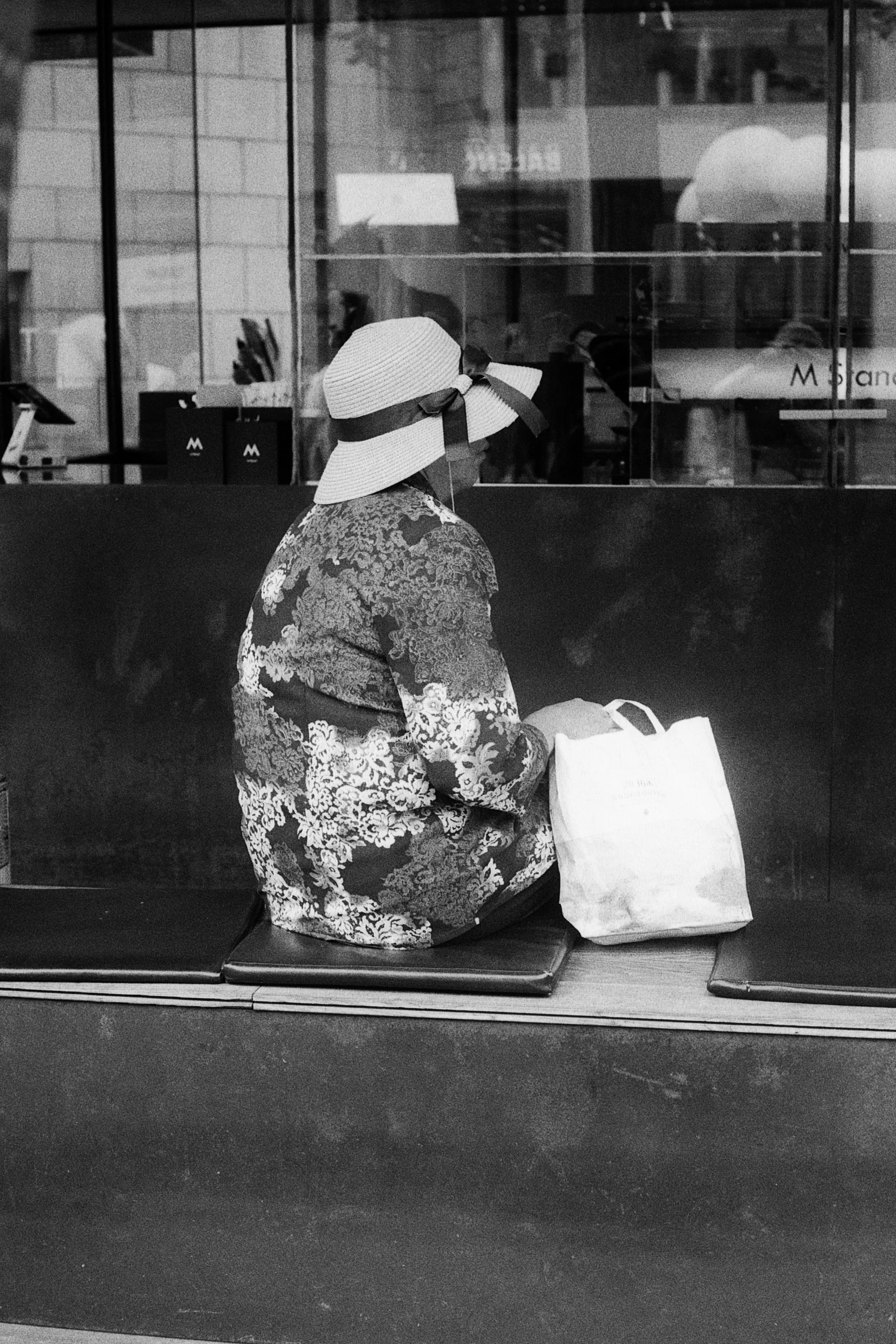 A person wearing a wide sun hat sits beside two bags in front of a reflective window.