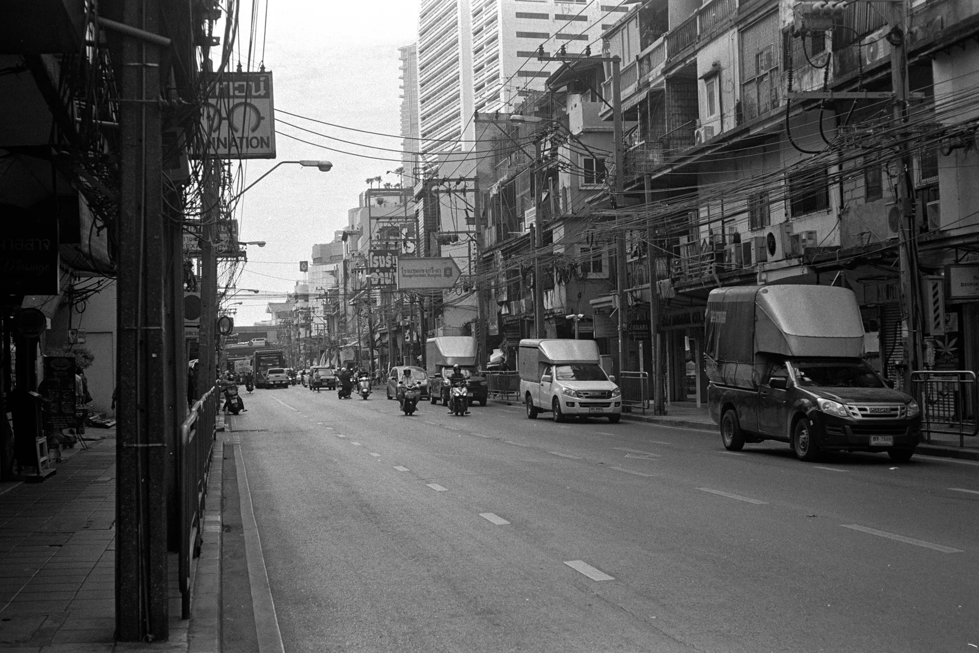 A broad street runs between rows of weathered buildings, parked cars, and overhead wires.