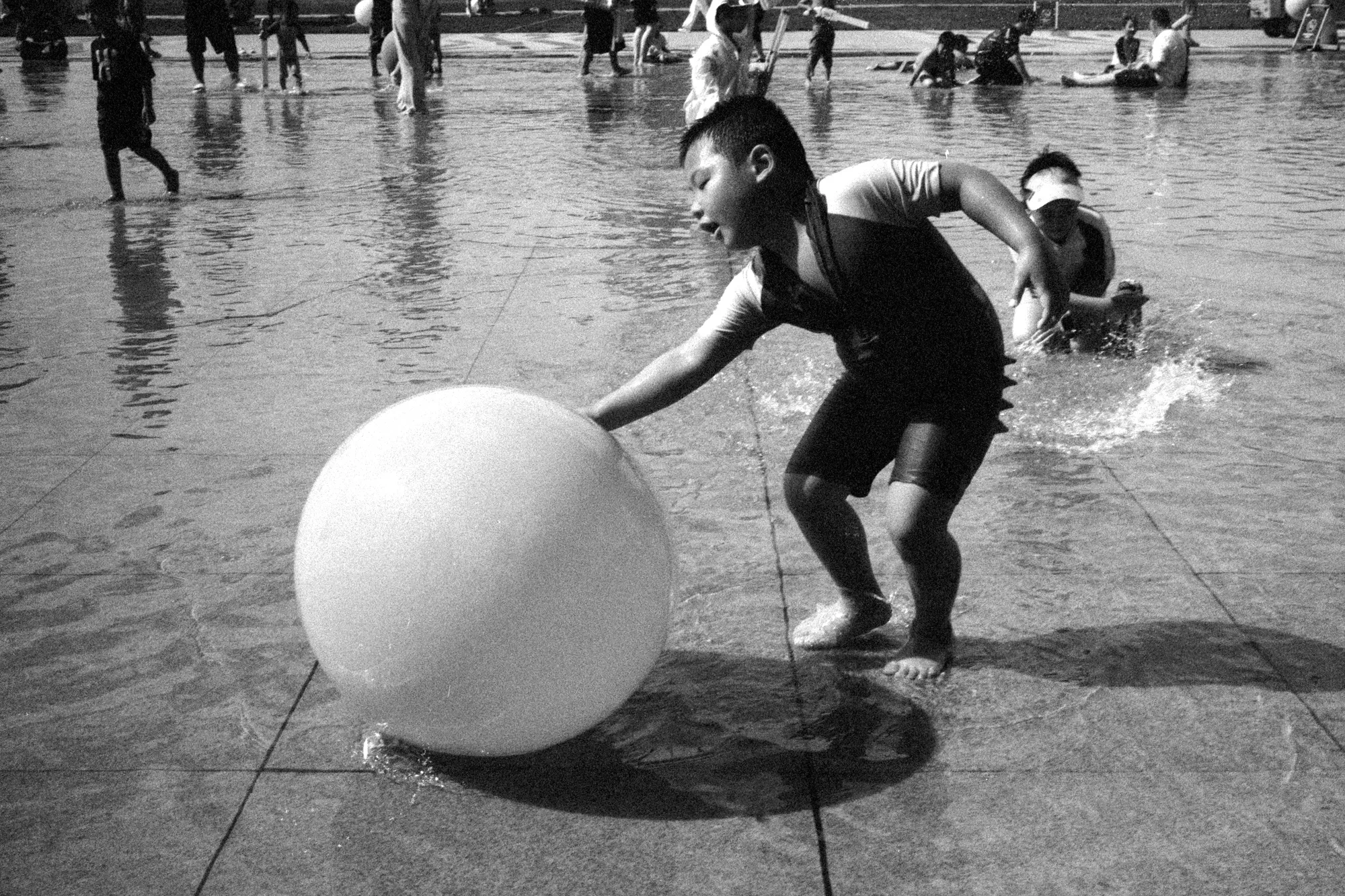 A child pushes a large white ball across a wet plaza while other children play behind him.