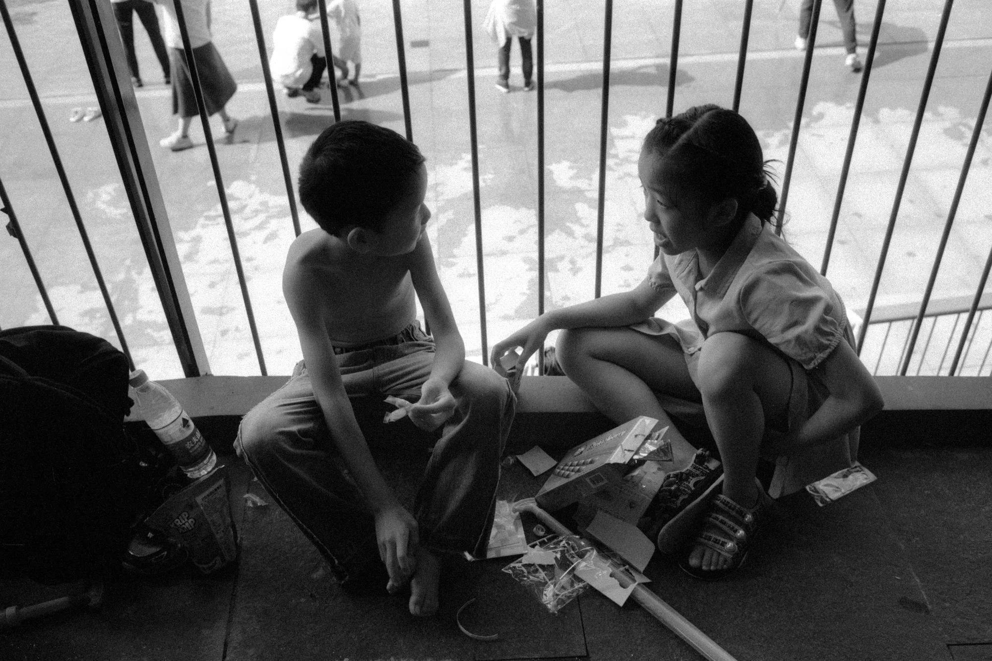 Two children sit beside a metal fence with books and papers spread around them.