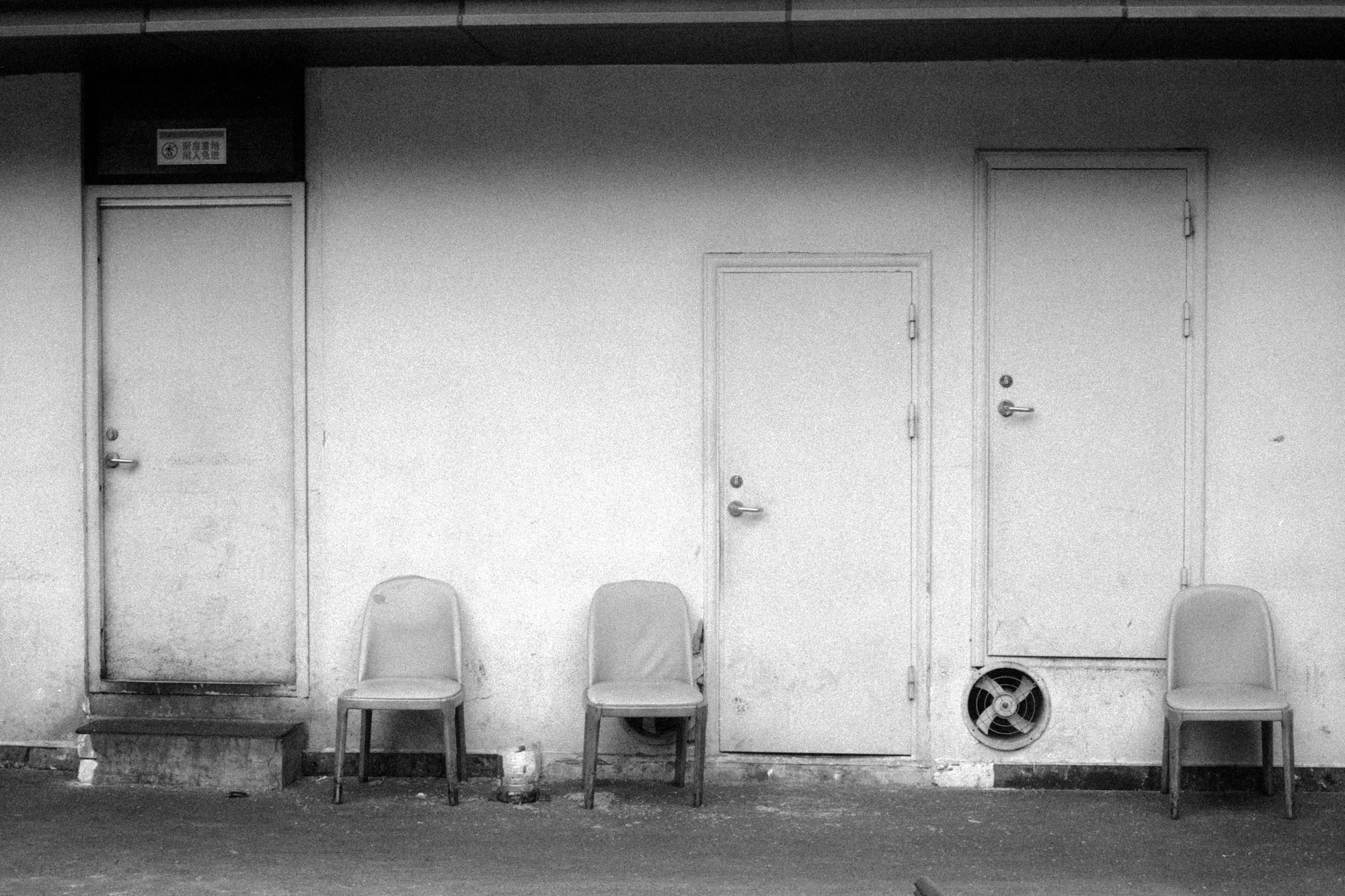 Three plastic chairs stand against a plain wall between two closed doors.